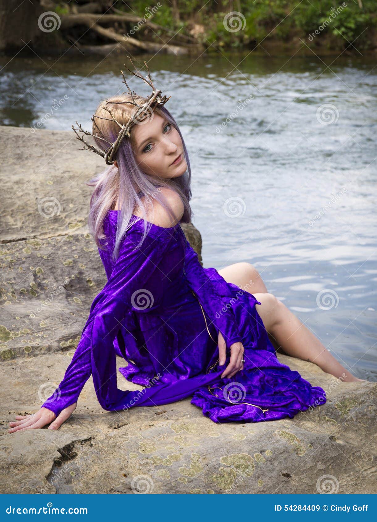 Young woman in purple gown sitting by a river. Beautiful young woman wearing purple velvet gown and twig crown sitting on some rocks by a river.