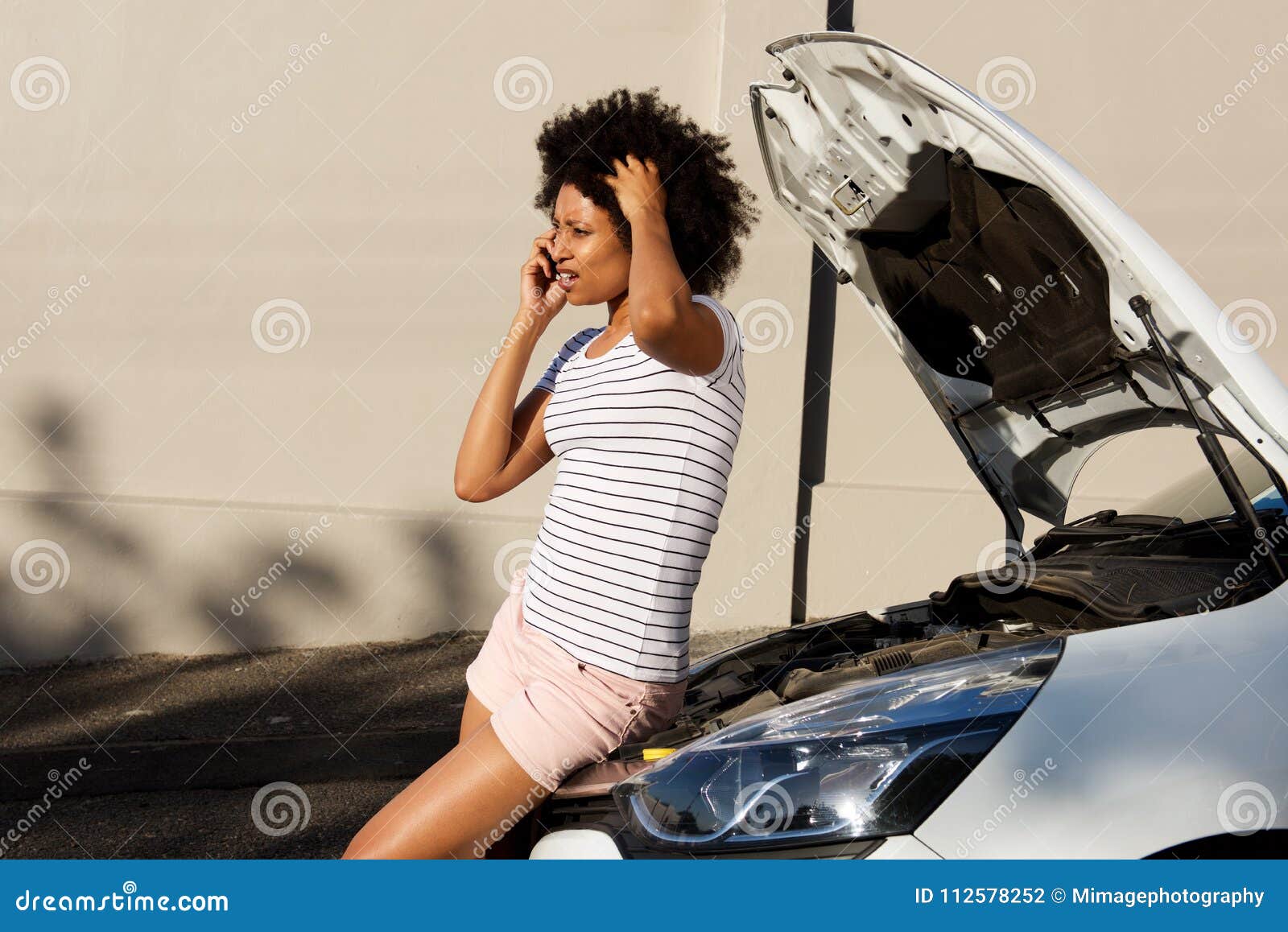 young woman standing by broken down car and making phone call for assistance