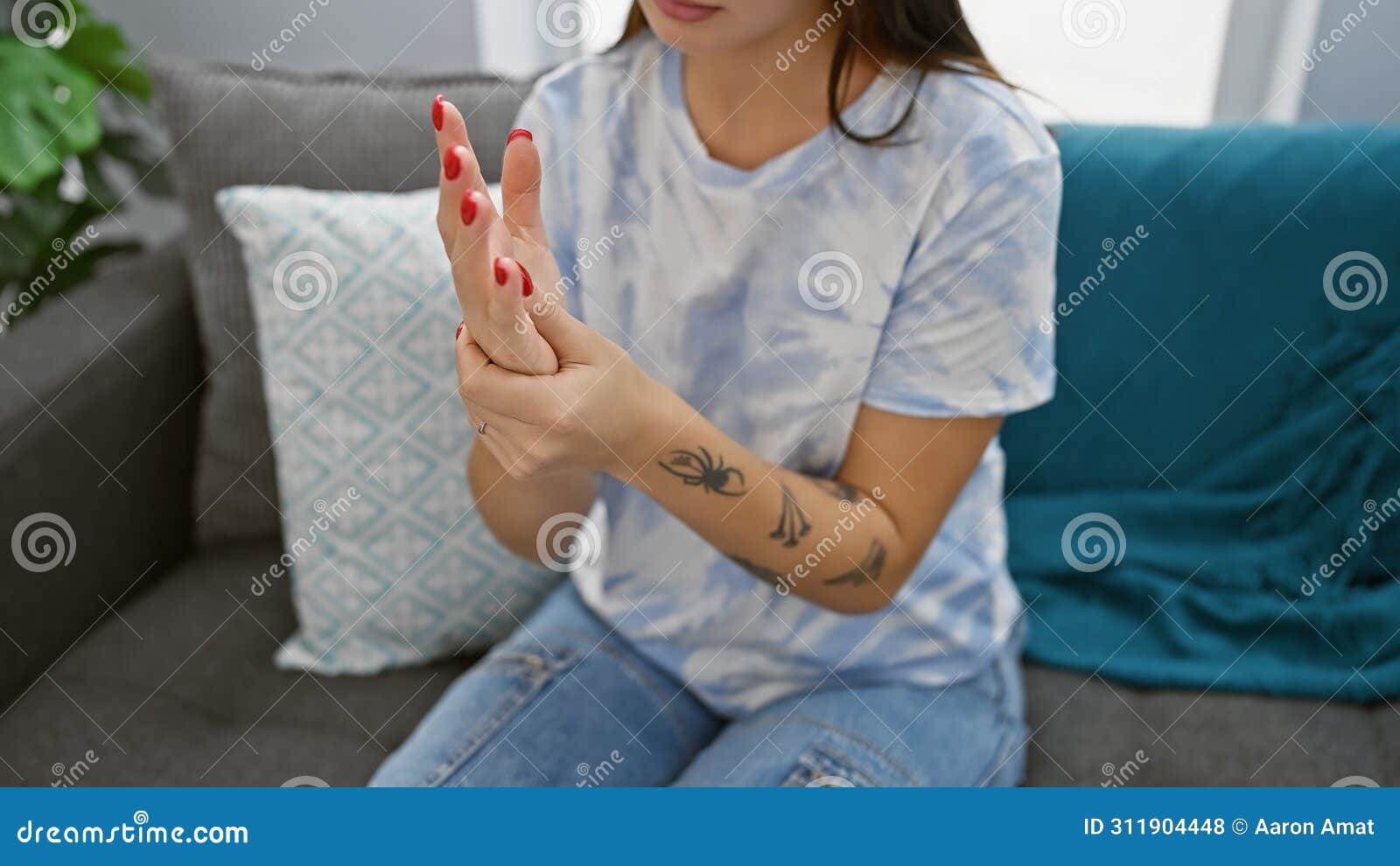 a young woman with a tattoo on her forearm massages her painful hand indoors on a couch