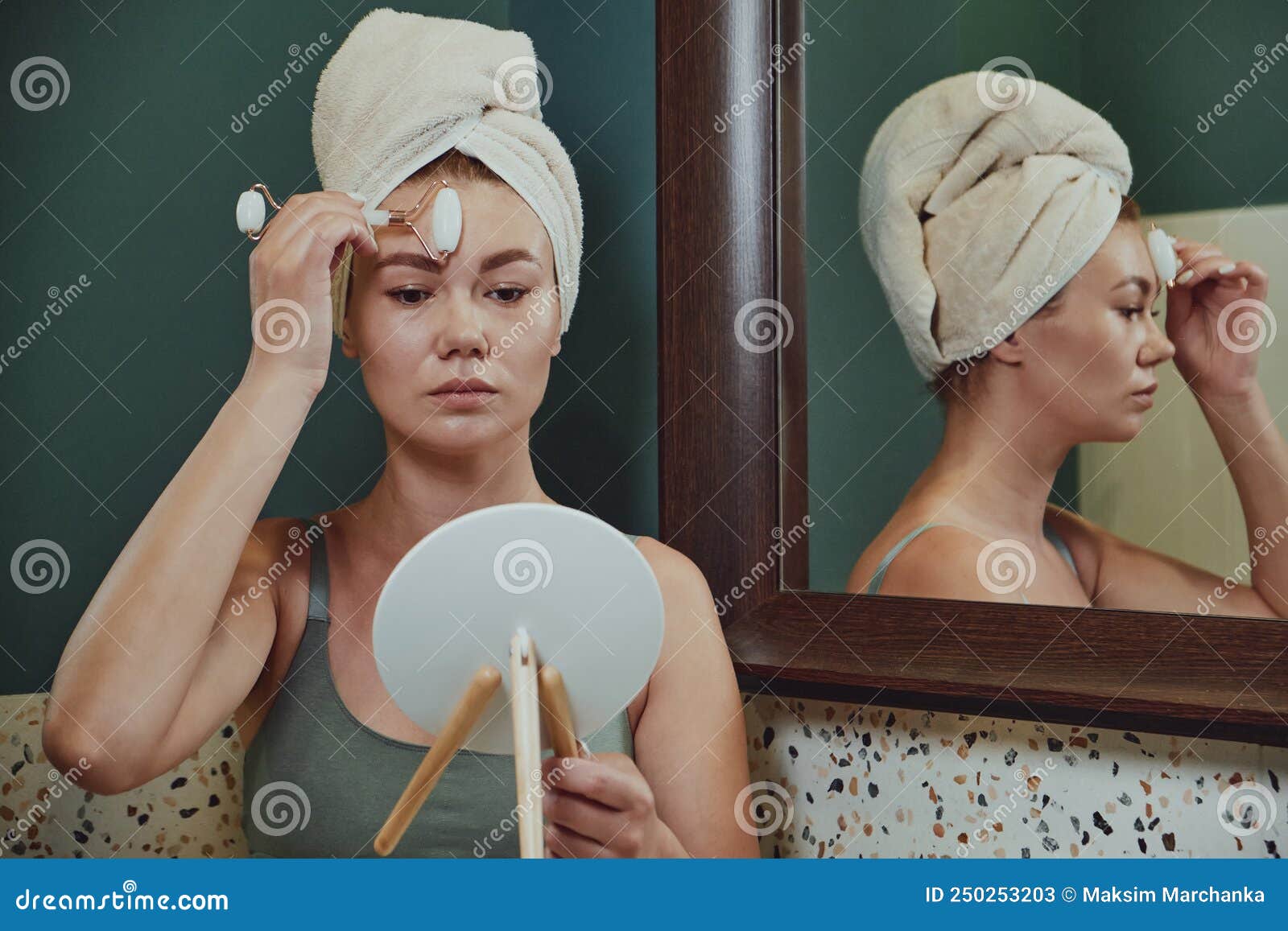 Young Woman Using Jade Facial Roller For Face Massage Sitting In Bathroom Looking In The Mirror