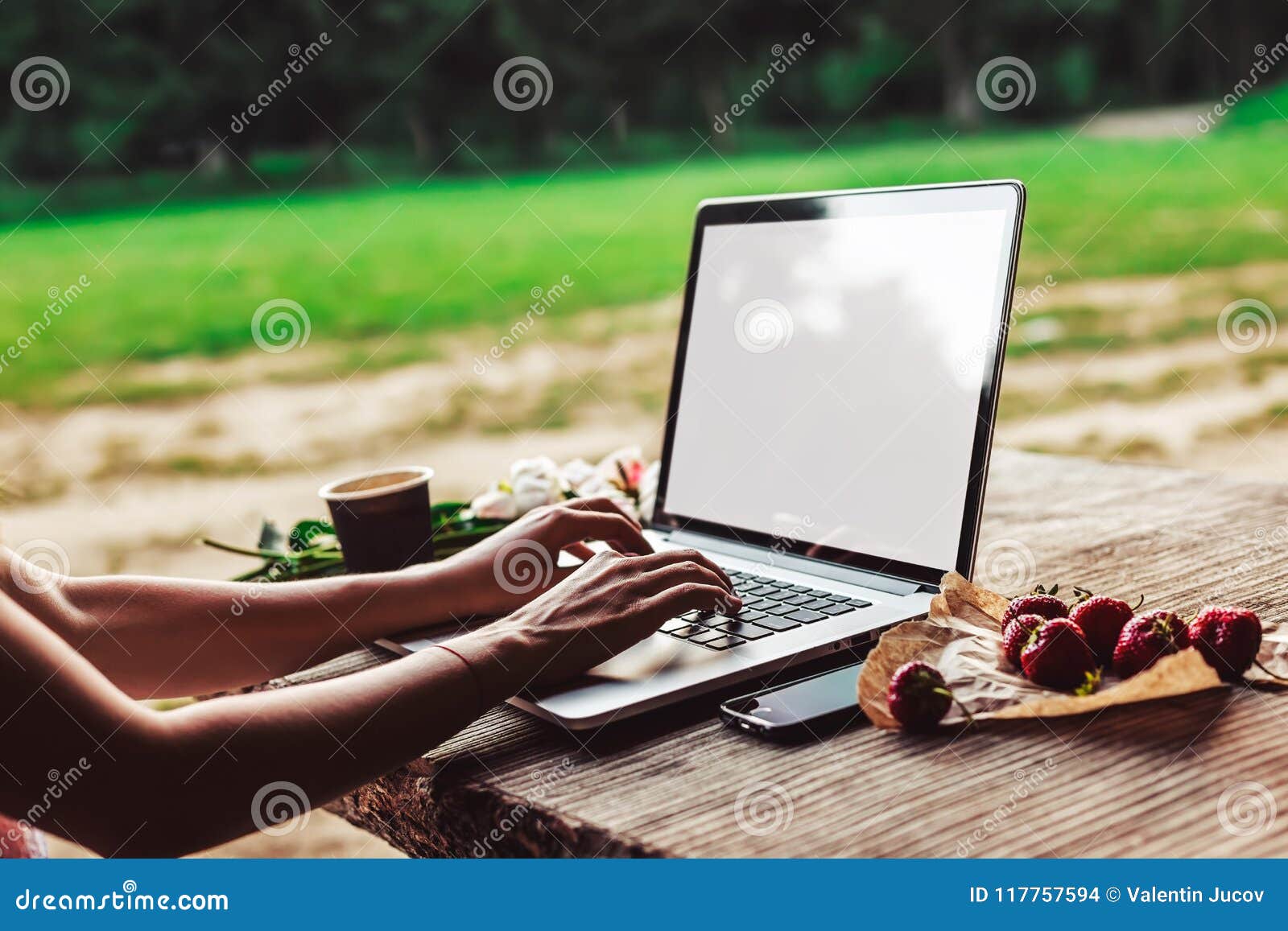 young woman using and typing laptop computer at rough wooden table with coffee cup, strawberries, bouquet of peonies flowers,