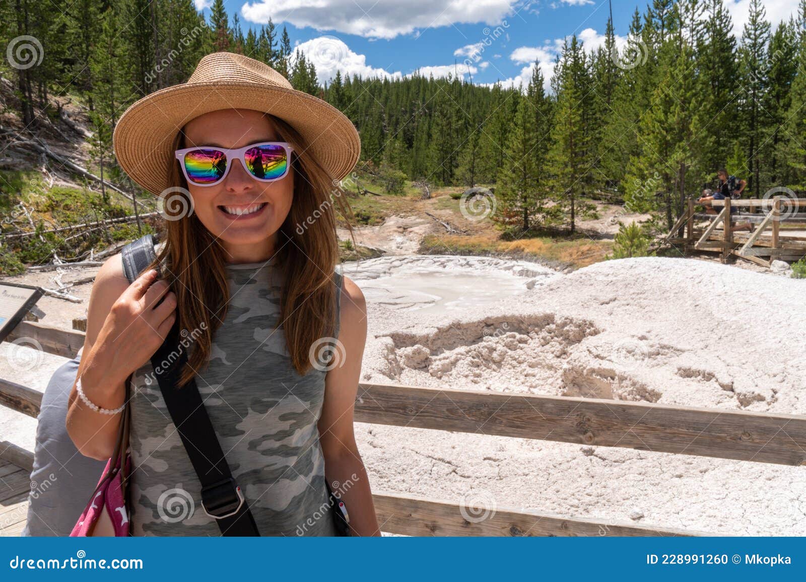 young woman wearing a camoflauge outfit and sun hat poses at the mud pots at artist paint pots in yellowstone