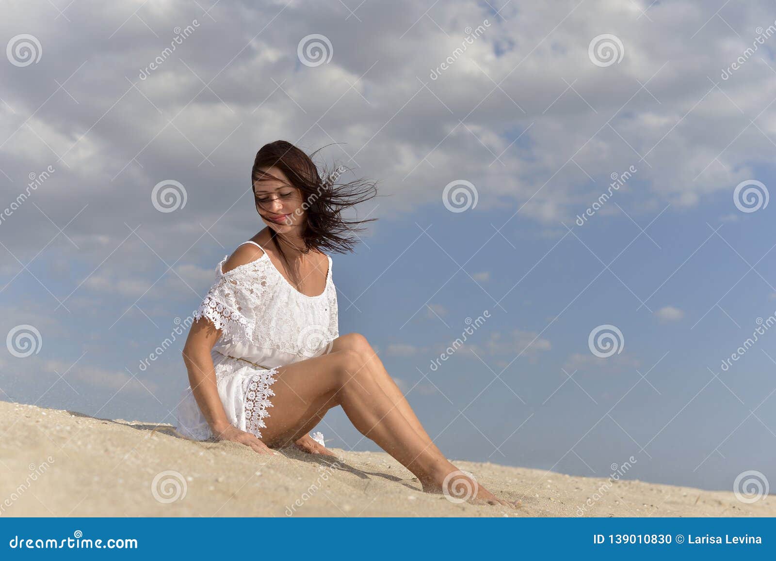 A Young Woman In A White Dress Sitting Barefoot On The Sandy Beach 