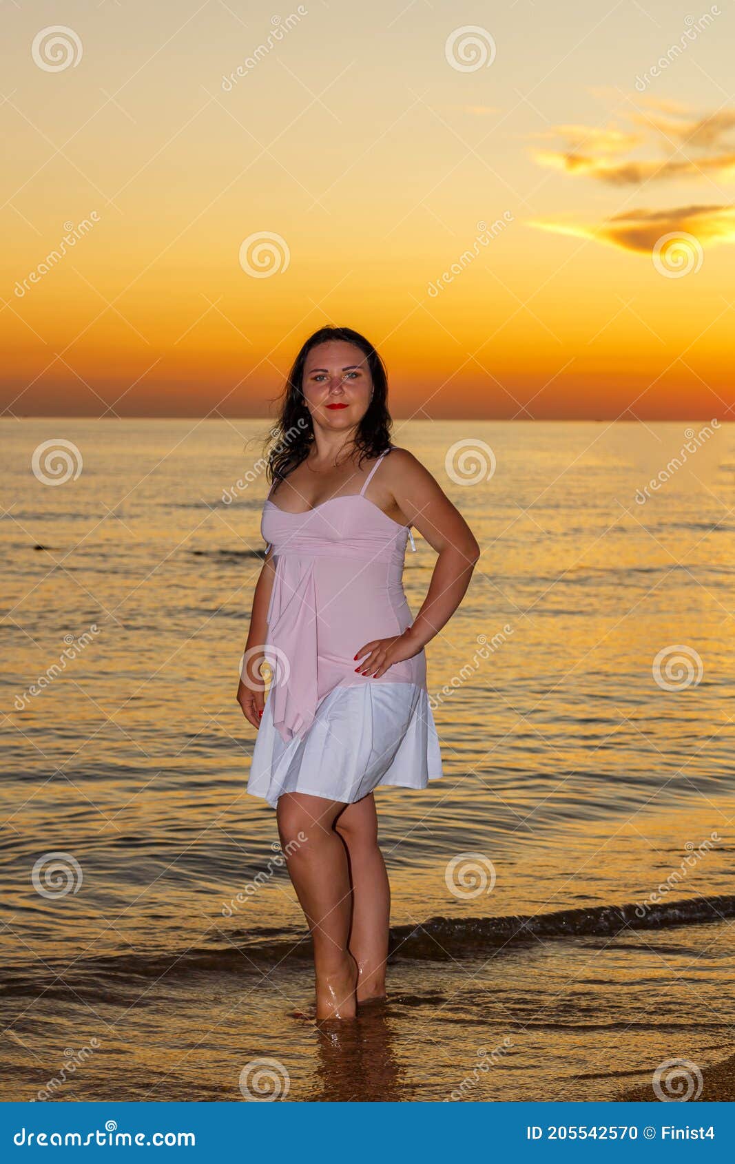 a young woman in a white dress stands on the seashore at sunset.
