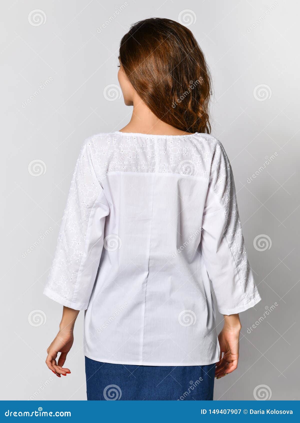 Back of a young brunette girl with long hair in a white blouse and blue skirt. on a light background.