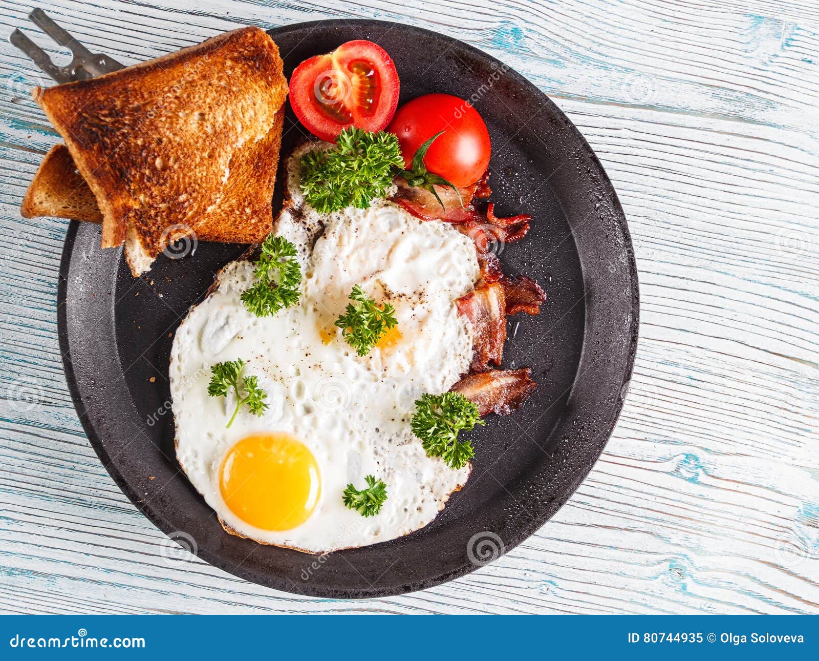 Äggfrukost med tomaten och persilja Selektivt fokusera. Ägg frukosterar med tomaten och persilja i järntappning som steker på träbakgrund Selektivt fokusera