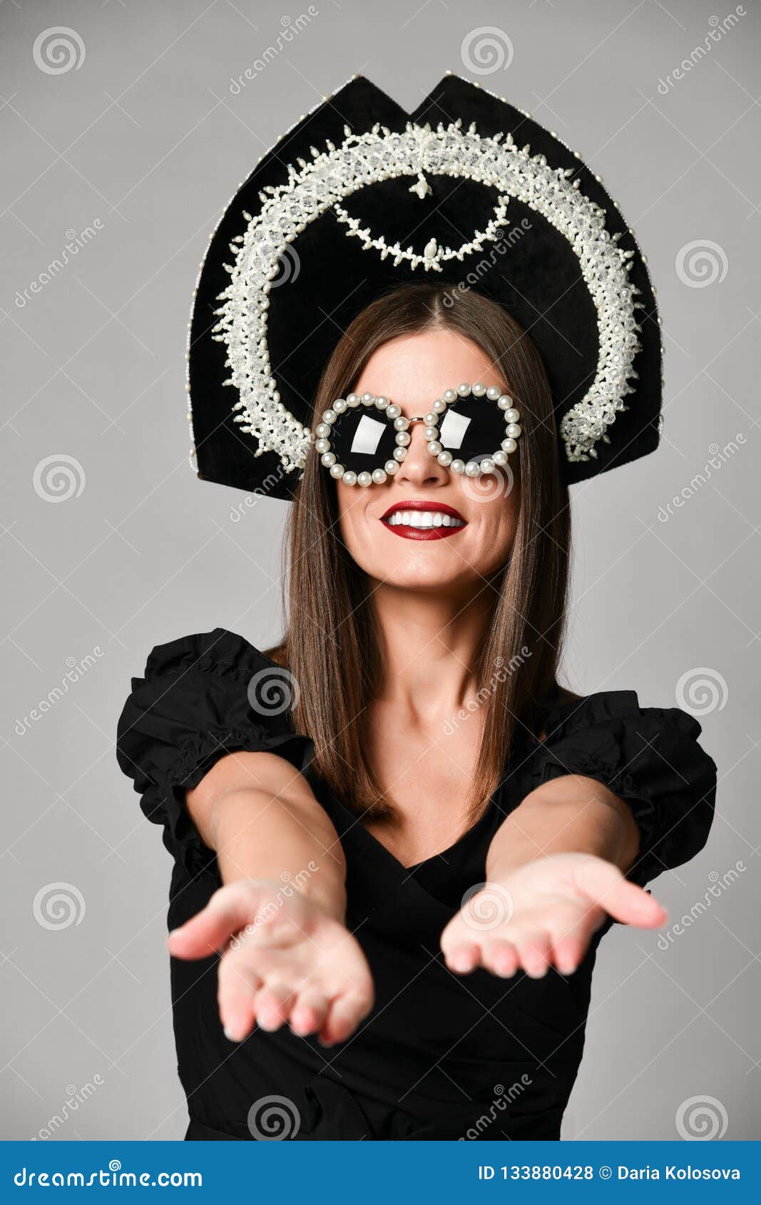 Élégance et type Portrait de studio de jeune femme magnifique dans peu de robe noire posant sur le fond gris. Élégance et type Le portrait de studio de la jeune femme magnifique dans peu de robe, lunettes de soleil et chapeau noirs de kokoshnik a étiré ses bras à la caméra en avant