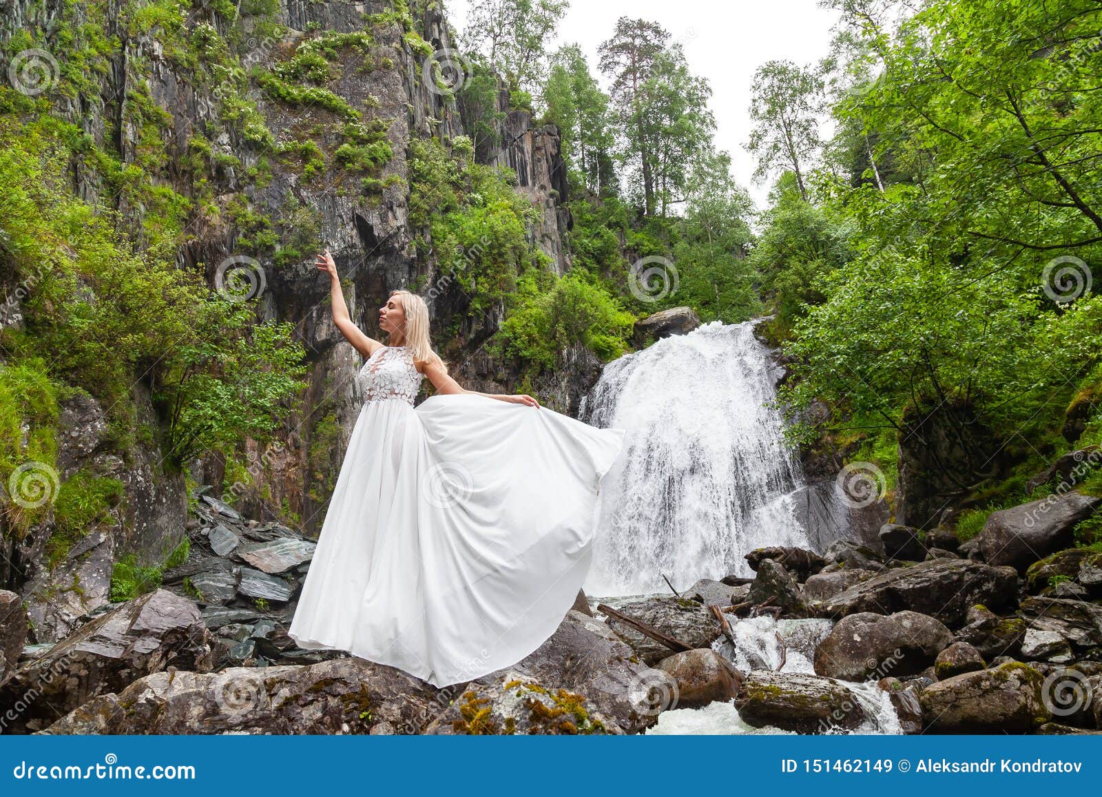 Ένα νέο ξανθό κορίτσι σε έναν κομψό θέτει σηκώνει ένα φόρεμα μπουντουάρ στα βουνά ενάντια σε έναν καταρράκτη και τις πέτρες αυξάνοντας τα χέρια της επάνω όπως ένα ballerina μια θερμή θερινή ημέρα σε Altai με τα πράσινα δέντρα