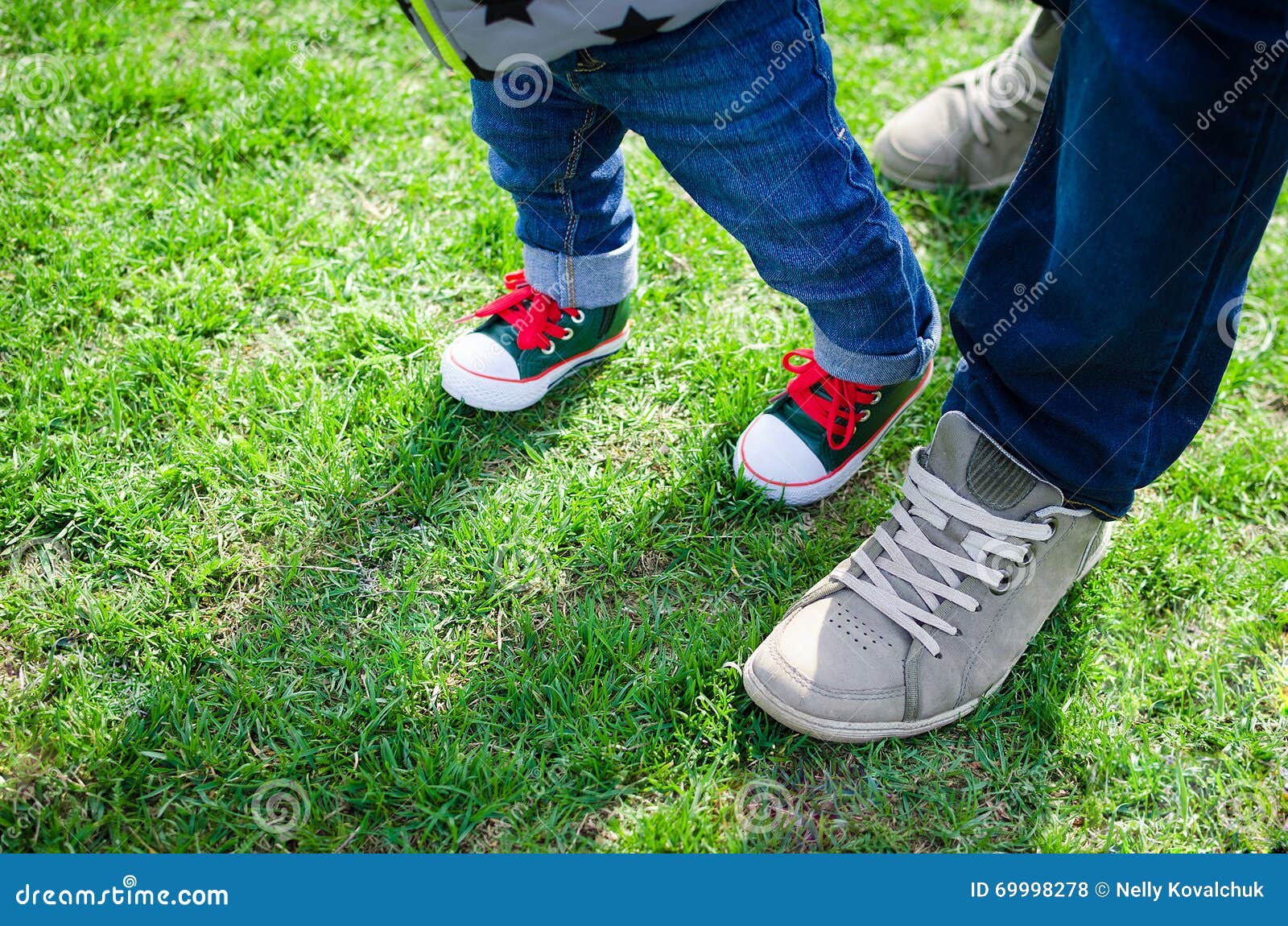 Έννοια μικρών παιδιών περπατήματος - λίγο αγόρι παιδιών μωρών που κάνει το χέρι του πρώτου βημάτων πατέρα εκμετάλλευσης πρώτα βήματα Πόδι του πατέρα και του γιου στα μοντέρνα πάνινα παπούτσια στο υπόβαθρο της πράσινης χλόης διάστημα αντιγράφων