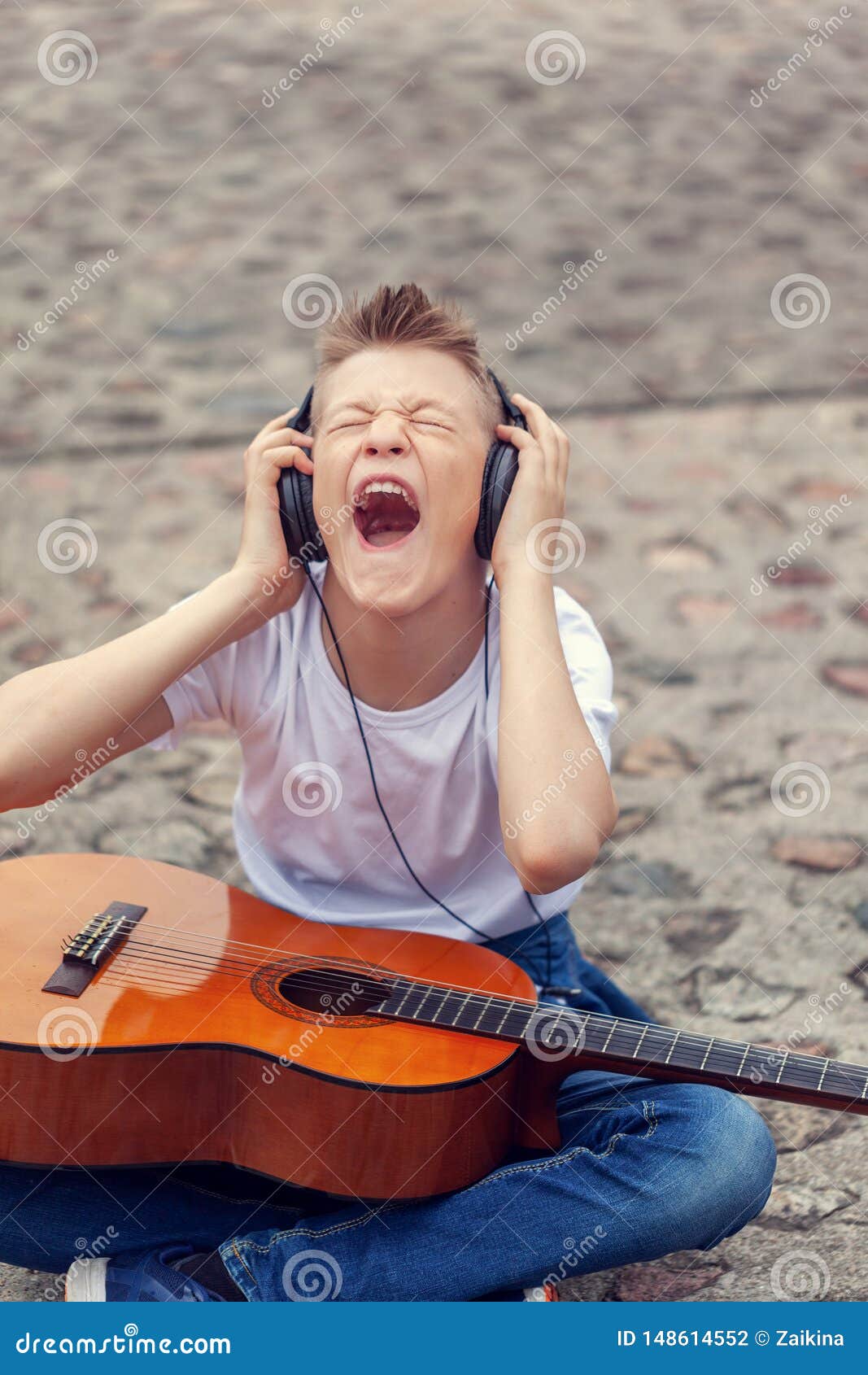 Έφηβος που ακούει τη μουσική στα ακουστικά και το τραγούδι κραυγής Συνεδρίαση νεαρών άνδρων με μια κιθάρα - υπαίθρια