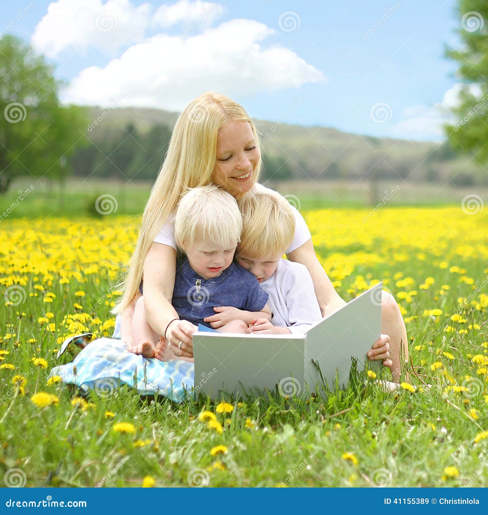 Βιβλίο ανάγνωσης μητέρων στα μικρά παιδιά έξω. Μια ευτυχής νέα μητέρα κάθεται έξω σε ένα λιβάδι λουλουδιών, διαβάζοντας ένα βιβλίο ιστορίας σε της στα μικρά παιδιά μια ηλιόλουστη θερινή ημέρα