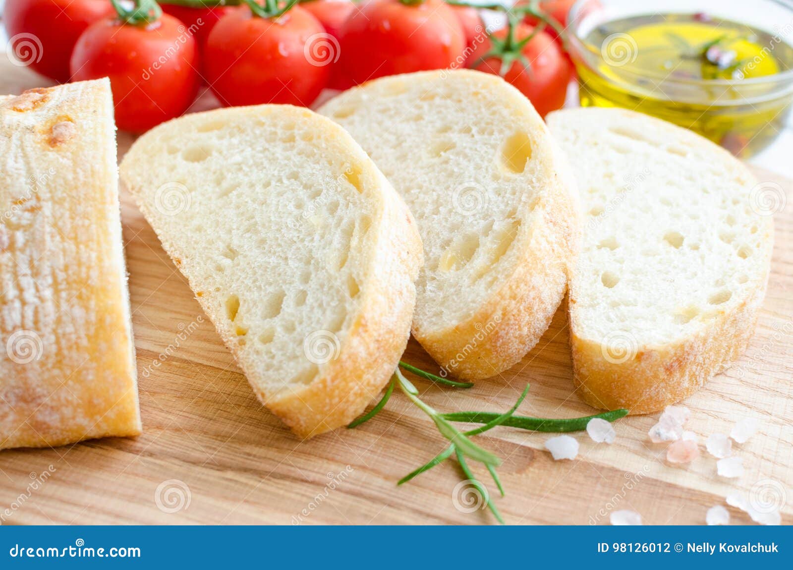 Ιταλικό ψωμί ciabatta. Τεμαχισμένο πρόσφατα ψημένο ψωμί ciabatta στον ξύλινο τέμνοντα πίνακα στον άσπρο αγροτικό πίνακα με το δεντρολίβανο, το άλας, τις ντομάτες και το ελαιόλαδο Ιταλική έννοια τροφίμων