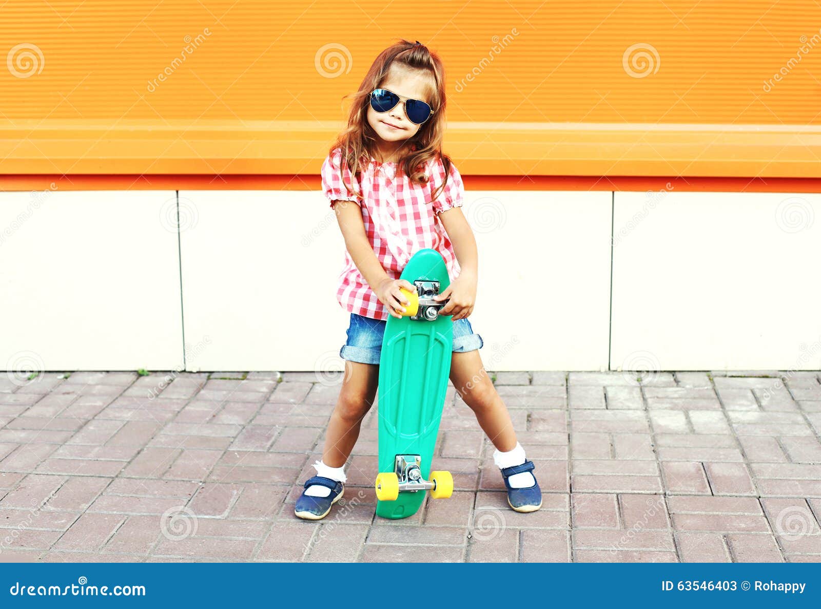 Μοντέρνο παιδί μικρών κοριτσιών μόδας με skateboard που φορά τα γυαλιά ηλίου και το ελεγμένο πουκάμισο στην πόλη