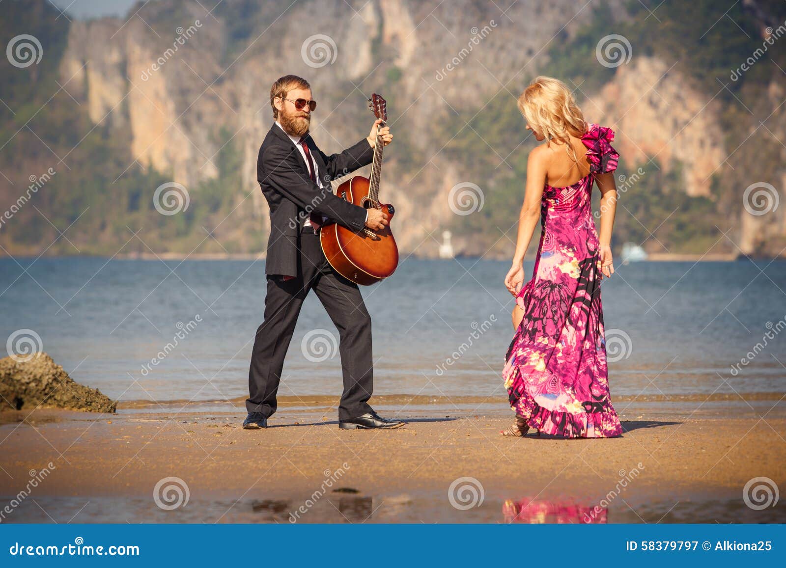 Πίσω-άποψη και κιθαρίστας χορού ξανθή στην παραλία. Χορεύοντας ξανθό κορίτσι κατά την υψηλή πίσω-άποψη παπουτσιών τακουνιών και γενειοφόρος κιθαρίστας στην παραλία at low tide ενάντια στη θάλασσα και το νησί