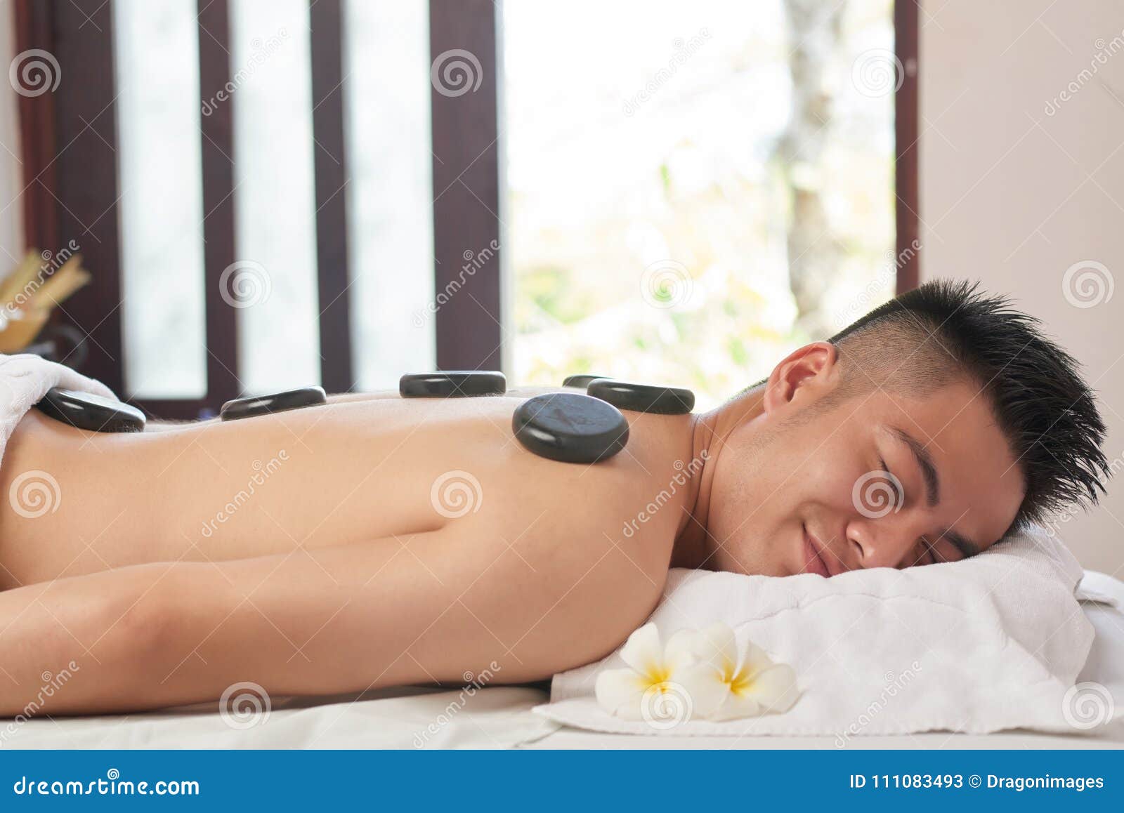 Азиатский человек на роскошном салоне курорта. Профилируйте взгляд усмехаясь азиатского человека лежа на таблице обработки с закрытыми глазами пока получающ горячий каменный массаж на роскошном салоне курорта