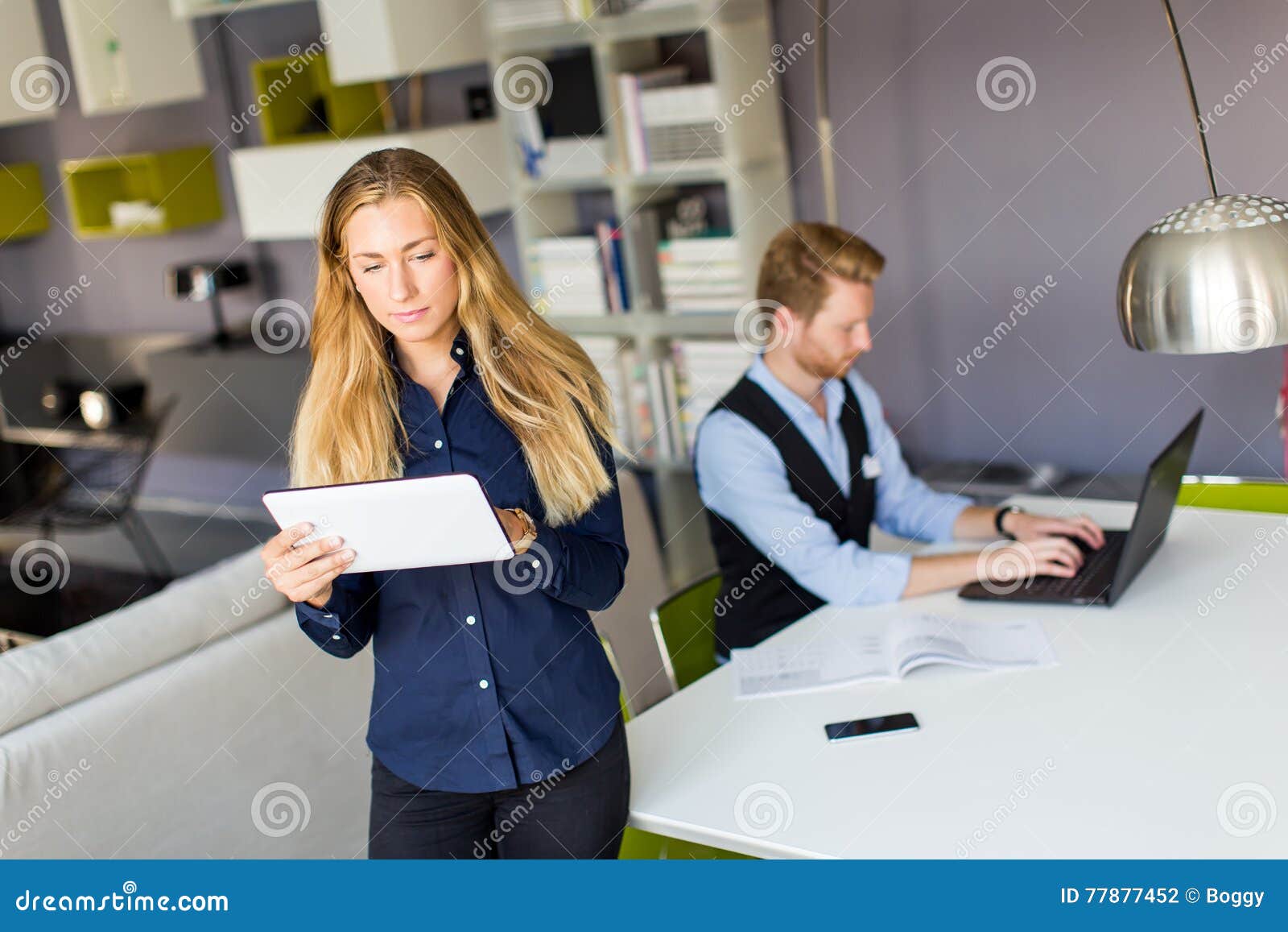 белизна офиса жизни фонового изображения 3d. Молодые бизнес-леди стоя в офисе и владениях таблетка пока бизнесмены работая на его компьтер-книжке на заднем плане