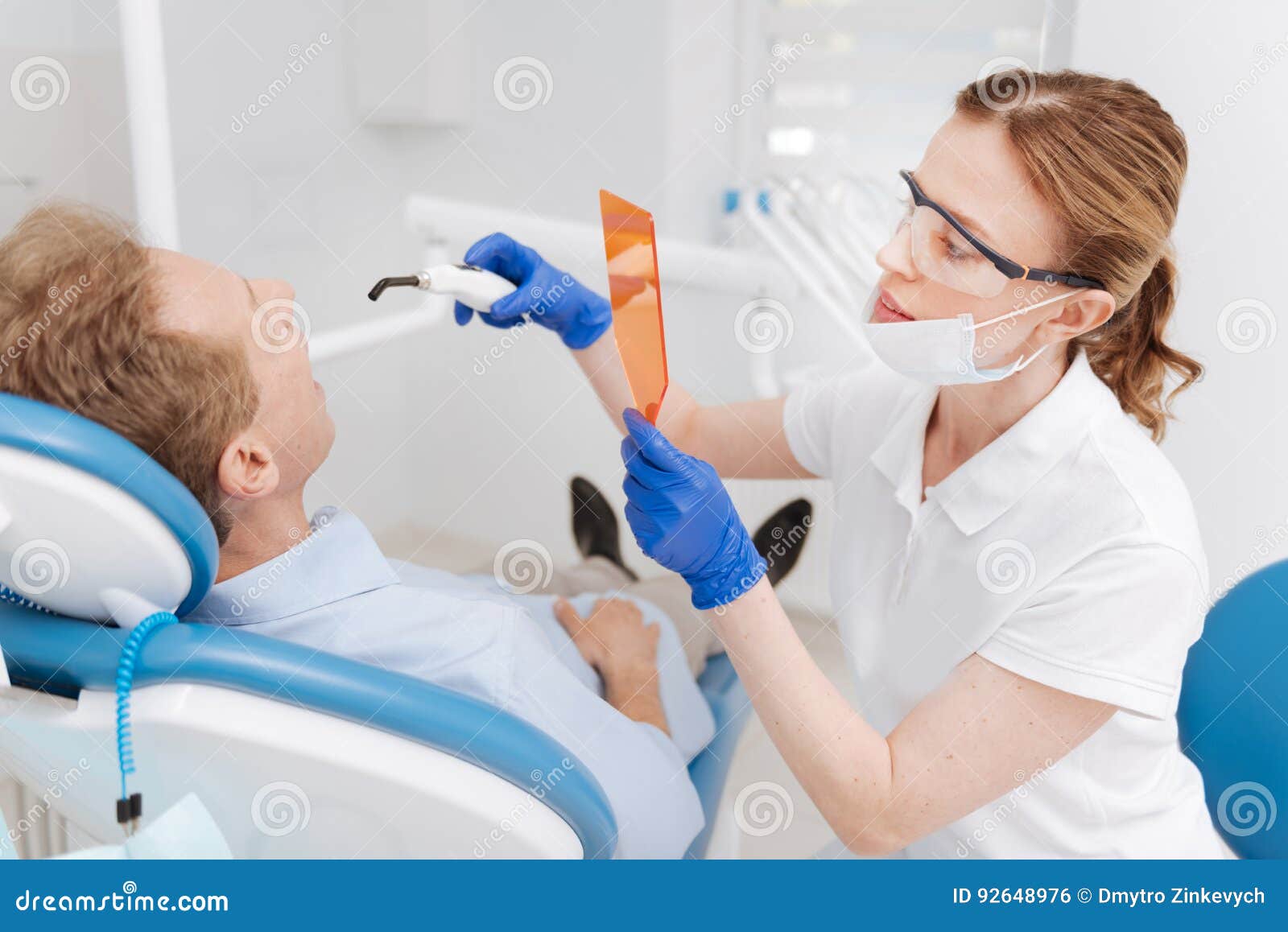 Видно скрупулезный дантист забеливая зубы пациентов. улыбка hollywood Тщательный тщательный квалифицированный доктор используя специальные приборы для защищать ее глаза и делать зубы людей смотря белый