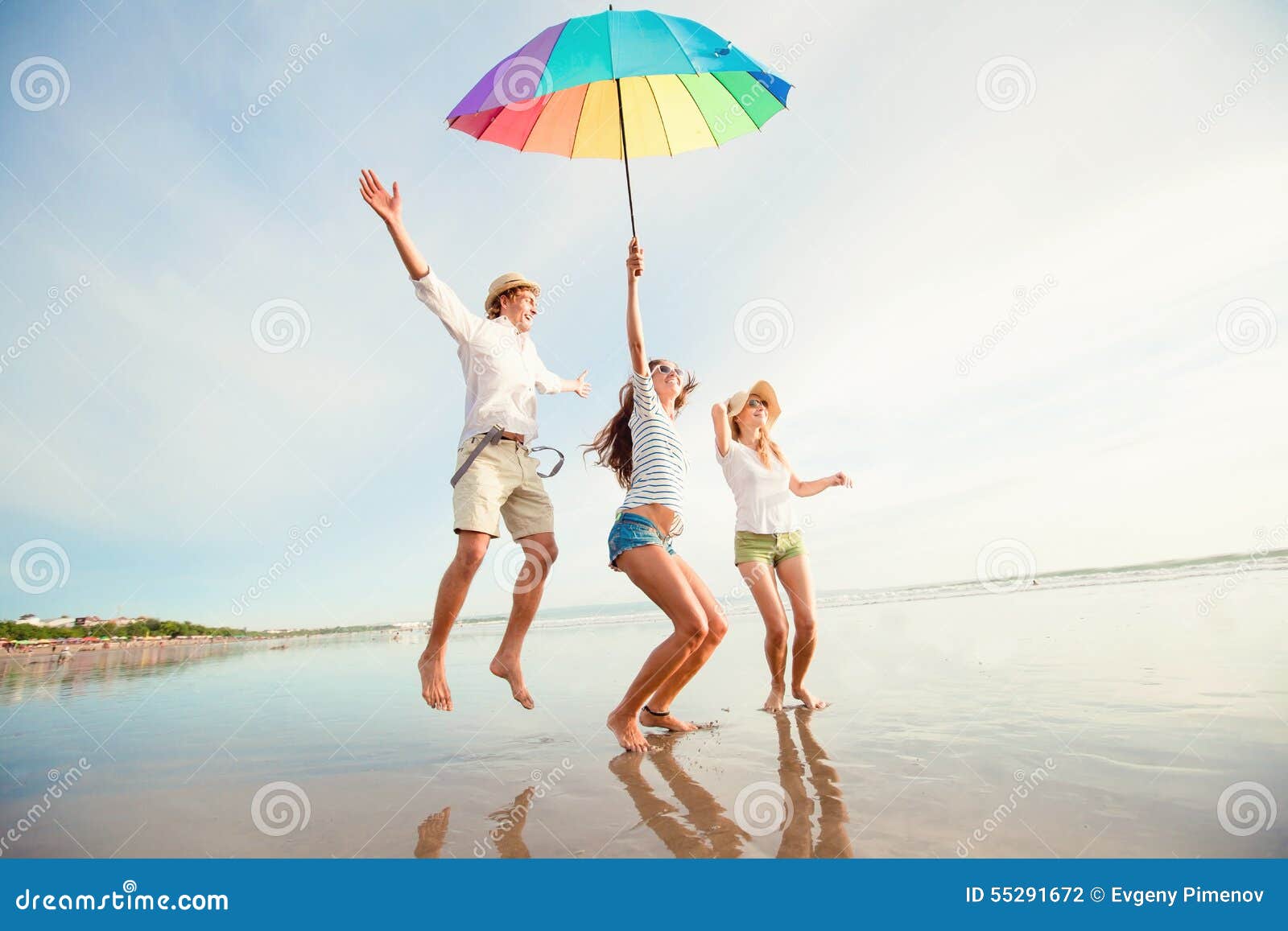 Группа в составе счастливое молодые люди имея потеху на. Группа в составе счастливое молодые люди имея потеху скача с зонтиком радуги на пляже на Бали