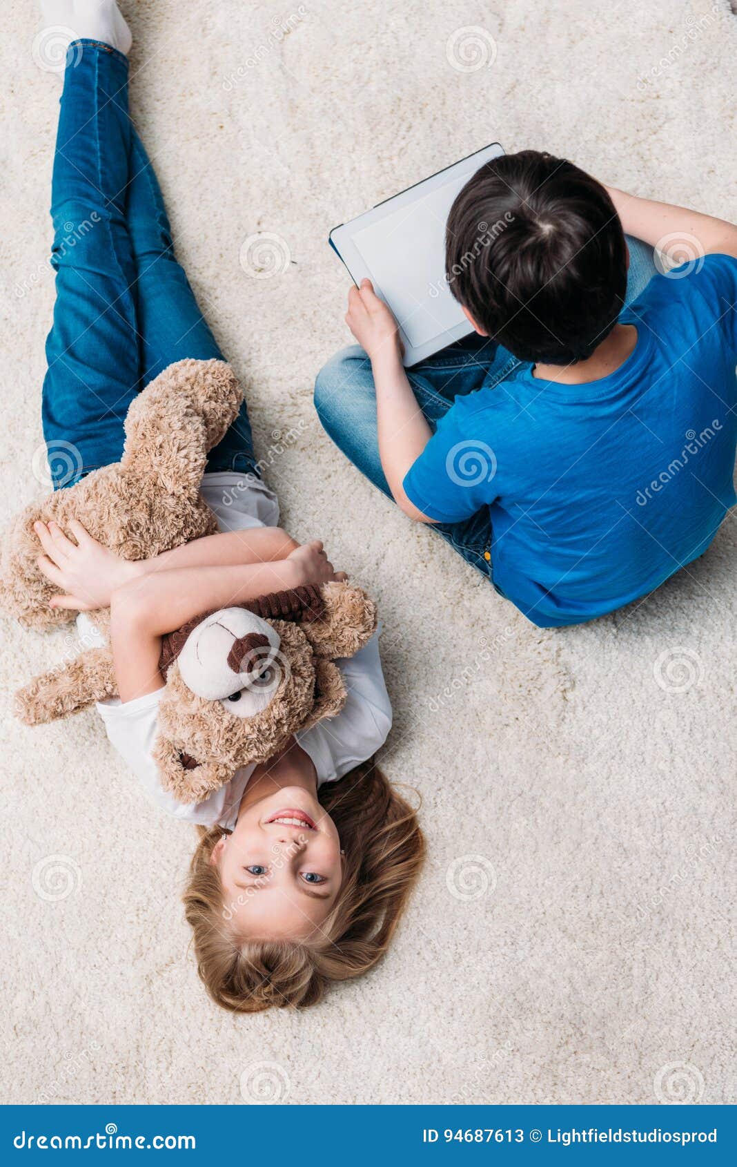 Девушка с плюшевым медвежонком и мальчиком с цифровой таблеткой на ковре дома. Маленькая девочка с плюшевым медвежонком и мальчиком с цифровой таблеткой на ковре дома