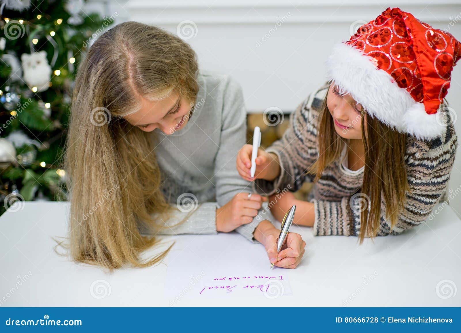 2 девушки пишут письмо к Санта Клаусу. 2 девушки в красной шляпе пишут письмо к Санта Клаусу на таблице около рождественской елки