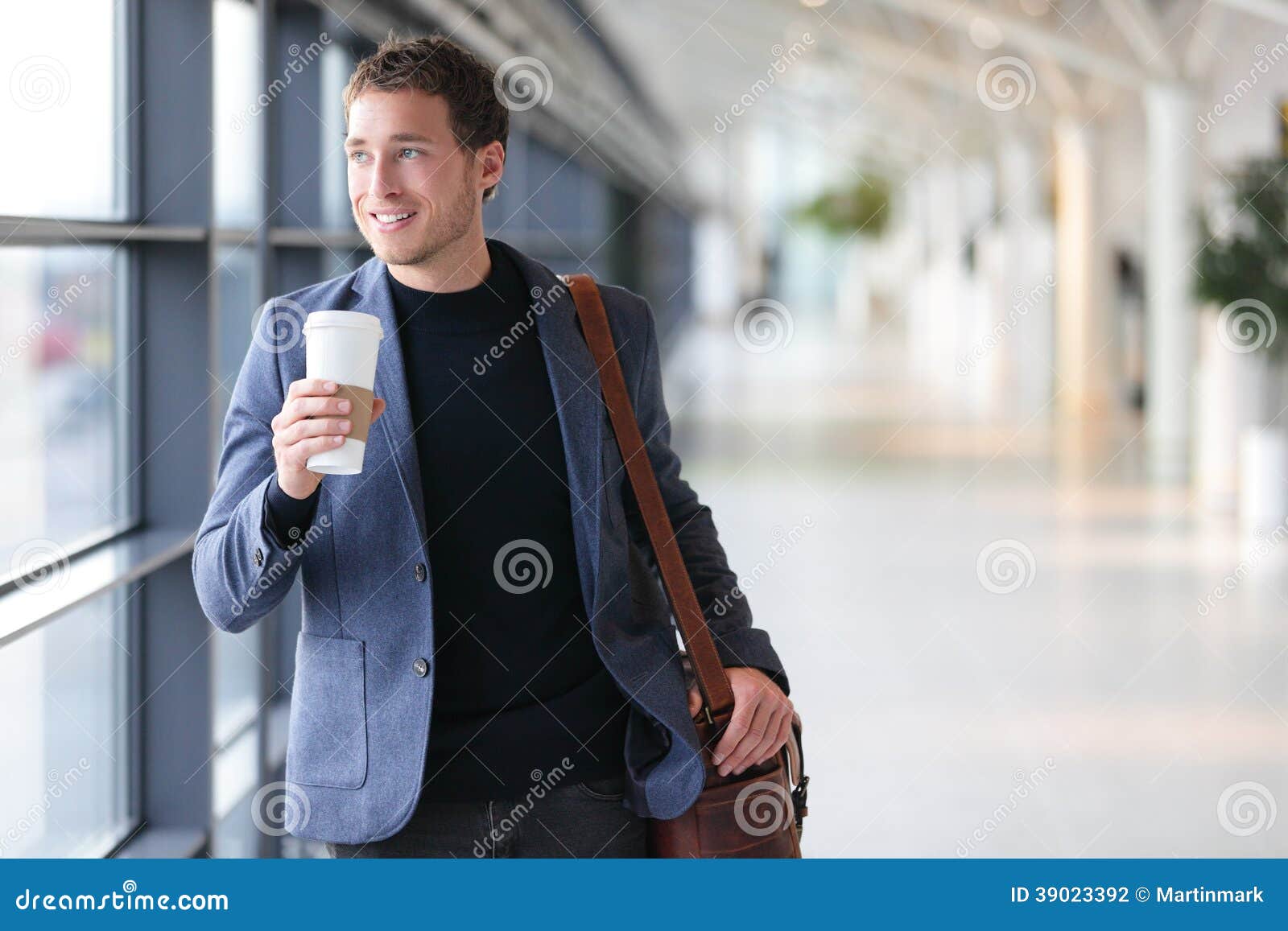 Кофе бизнесмена выпивая идя в авиапорт. Куртка костюма вскользь городского профессионала усмехаясь счастливая нося держа устранимую кофейную чашку на перемещении. Красивая мужская модель в его двадчадках.
