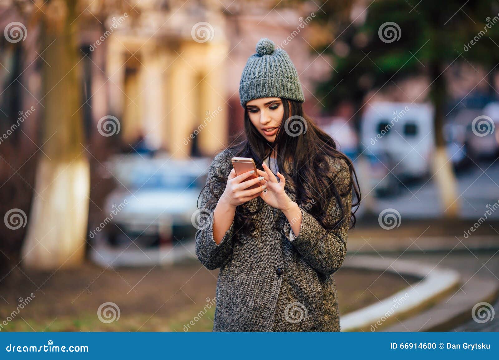 Молодая красивая вскользь девушка вызывая на ее сотовом телефоне на улице города весны. Молодая красивая вскользь девушка вызывая на ее сотовом телефоне в куртке и шляпа усмехаются на улице города весны Взгляд улицы весны