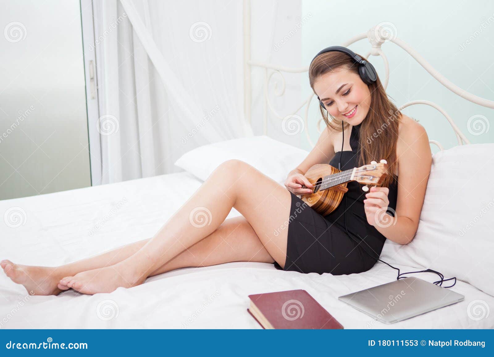 Молодая женщина сидящая на кровати играет укулеле слушая музыку наушниками в спальне в