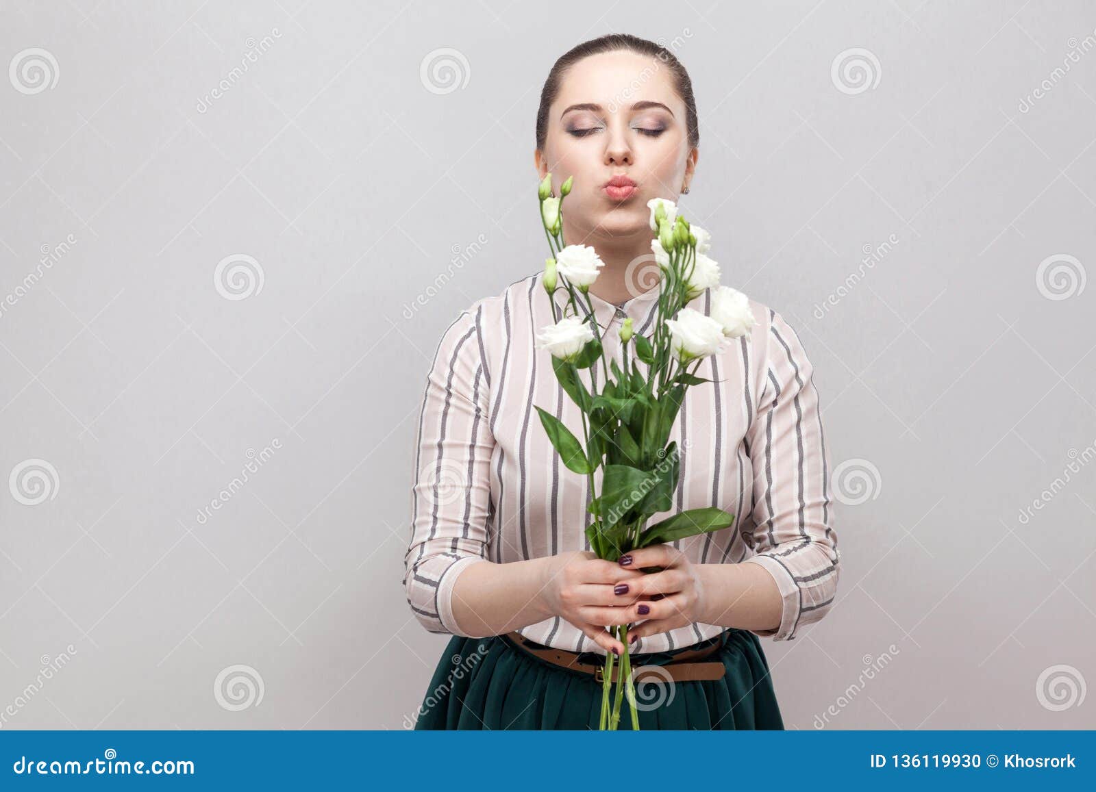 Портрет привлекательной романтичной прекрасной молодой женщины в striped рубашке и зеленый букет удерживания юбки белых цветков и отправки поцелуя воздуха Крытый, съемка студии, на серой предпосылке, космос экземпляра