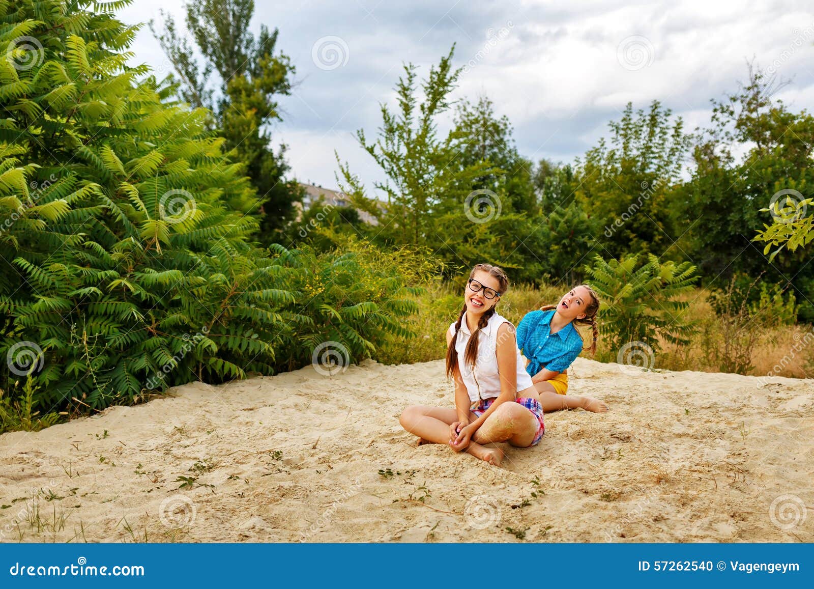 Самые лучшие подруги сидя на песке на пляже в лете паркуют Предназначенная для подростков девушка одела в шортах и рубашке На летних каникулах Концепция истинного приятельства