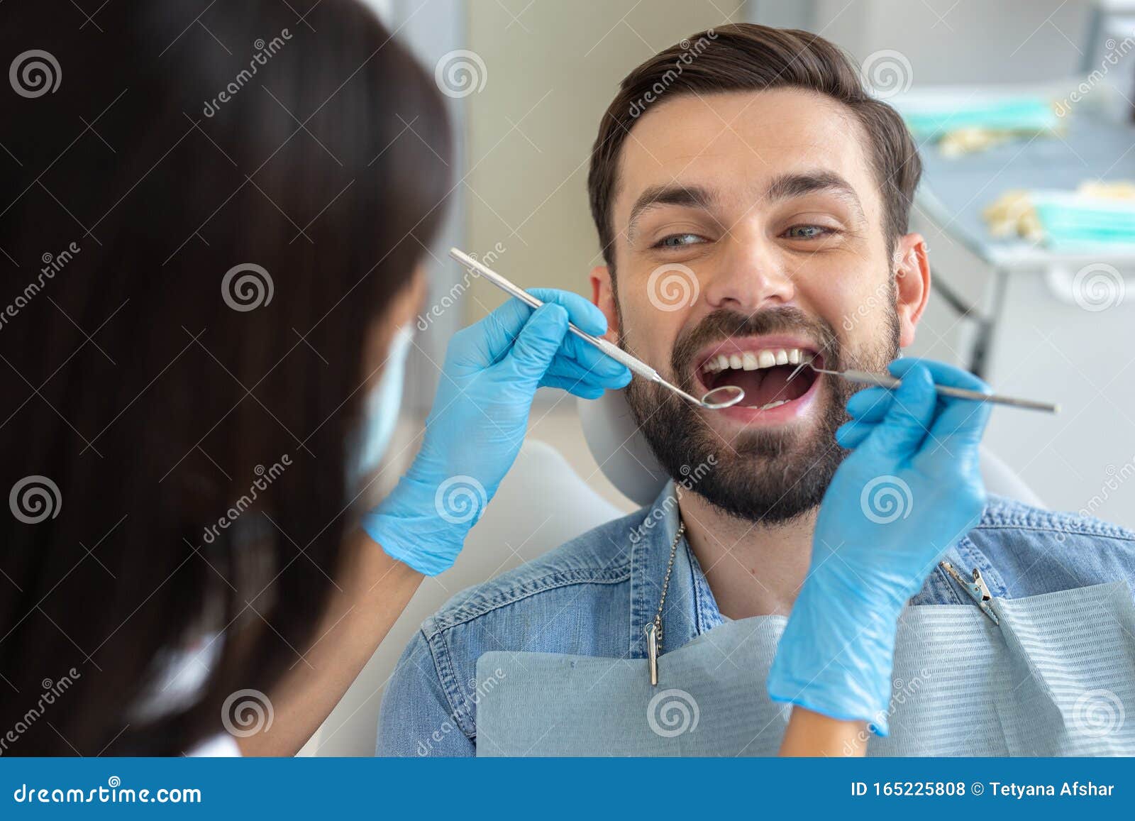 стоматолог изучает зубы симпатичного улыбающегося клиента в ...