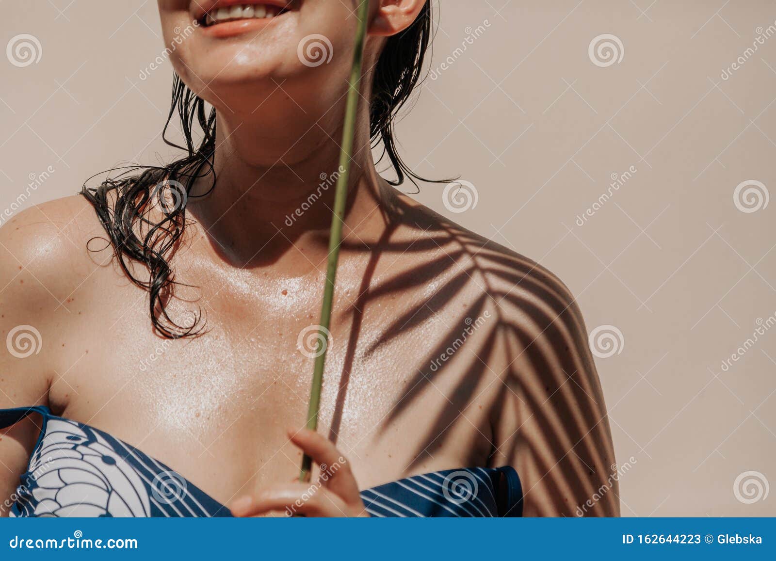 Тень ладони на теле девушки в купальнике. Загоревшая девушка в купальнике стоит на светлом фоне На левом плече находится тень от ладони с узкими листьями