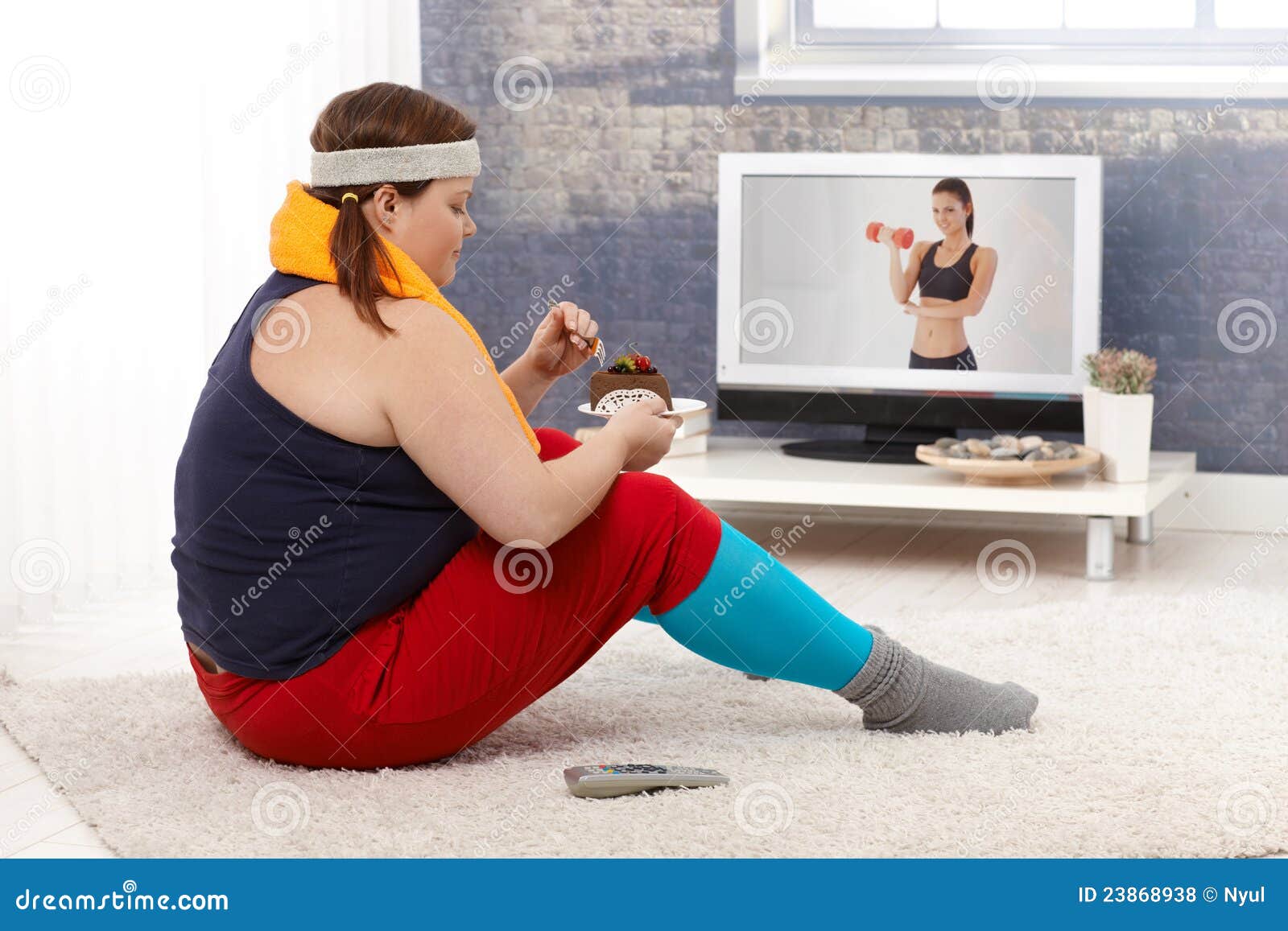 Тучная женщина есть торт шоколада в sportswear. Тучные женщины сидя на поле с тортом шоколада пока наблюдающ программу пригодности на телевидении.