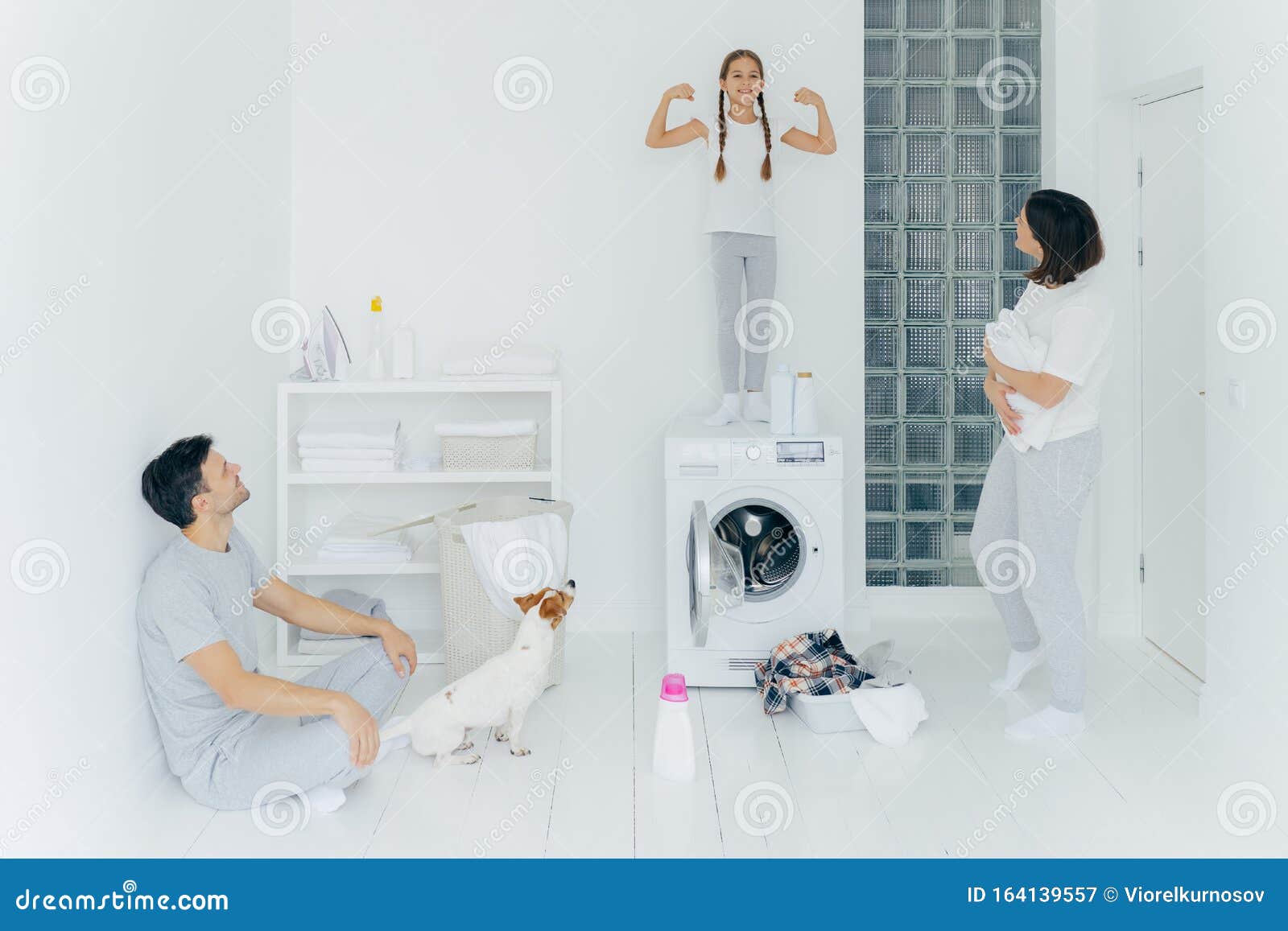 Фотография счастливых школьниц позирует на вершине стиральной машины, показывает мышцы, поднимает руки, готовая помочь родителям с стиркой или стиркой Женщины и мужчины позируют в стиральной комнате с собакой и маленьким ребенком