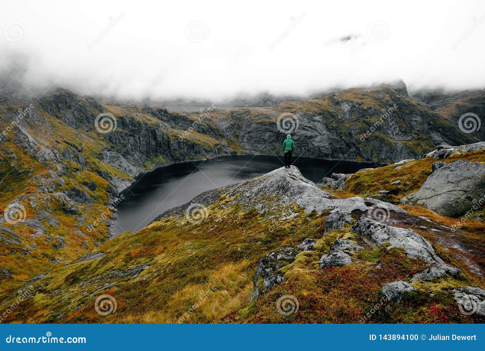 Человек на высокогорном озере, горной тропе Munken, островах Lofoten, Норвегии. Положение человека на крае высокогорного озера на горной тропе Munken около Sorvagen, Moskenesoy на островах Lofoten, Норвегии на туманном утре падения