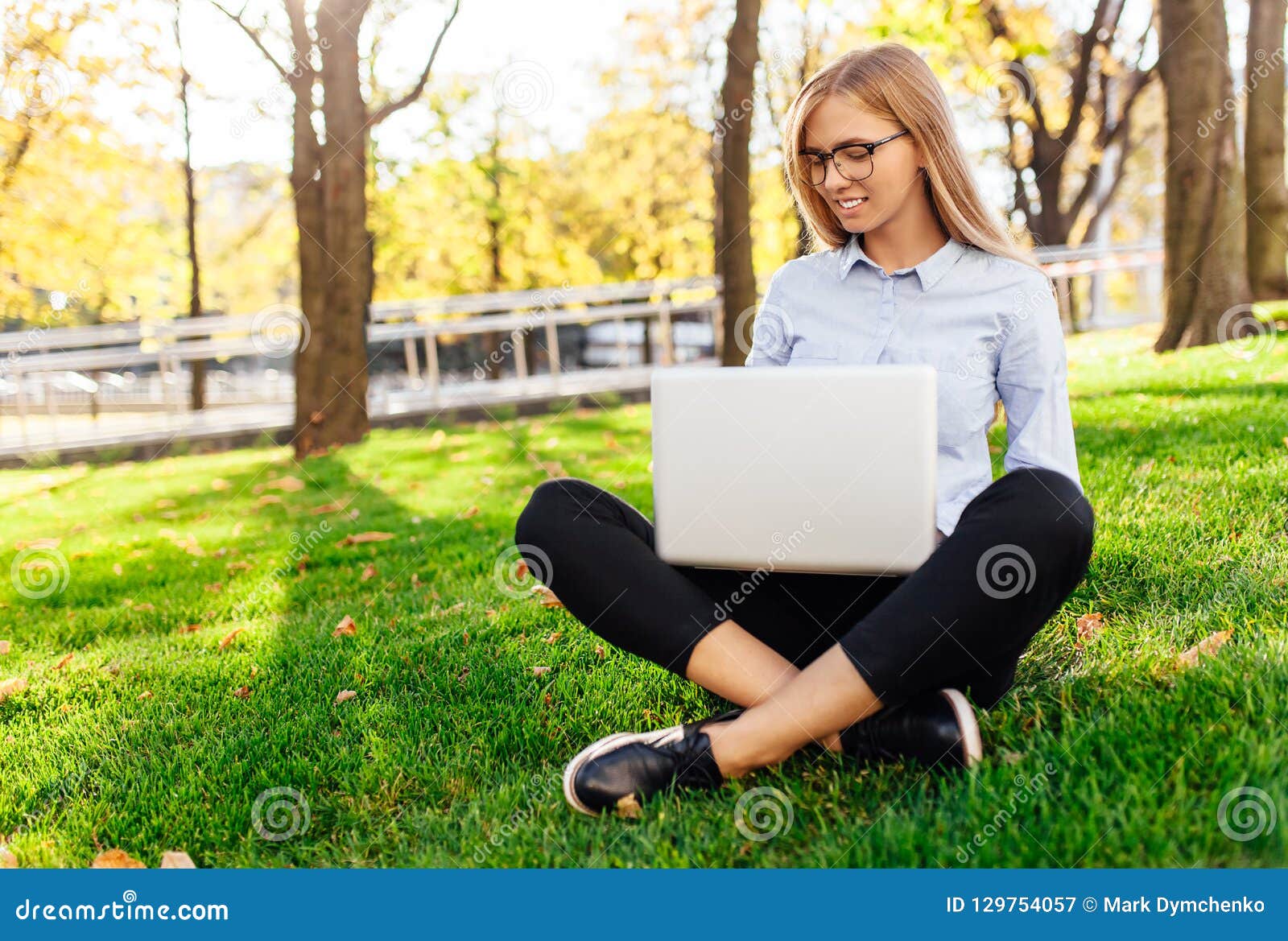 一个年轻惊人的夫人的图象，坐在公园，使用手提电脑，坐绿色草坪. 戴眼镜的年轻可爱的女孩，妇女坐草，研究一台膝上型计算机，在绿色草坪户外的一个城市公园 自由职业者的企业概念