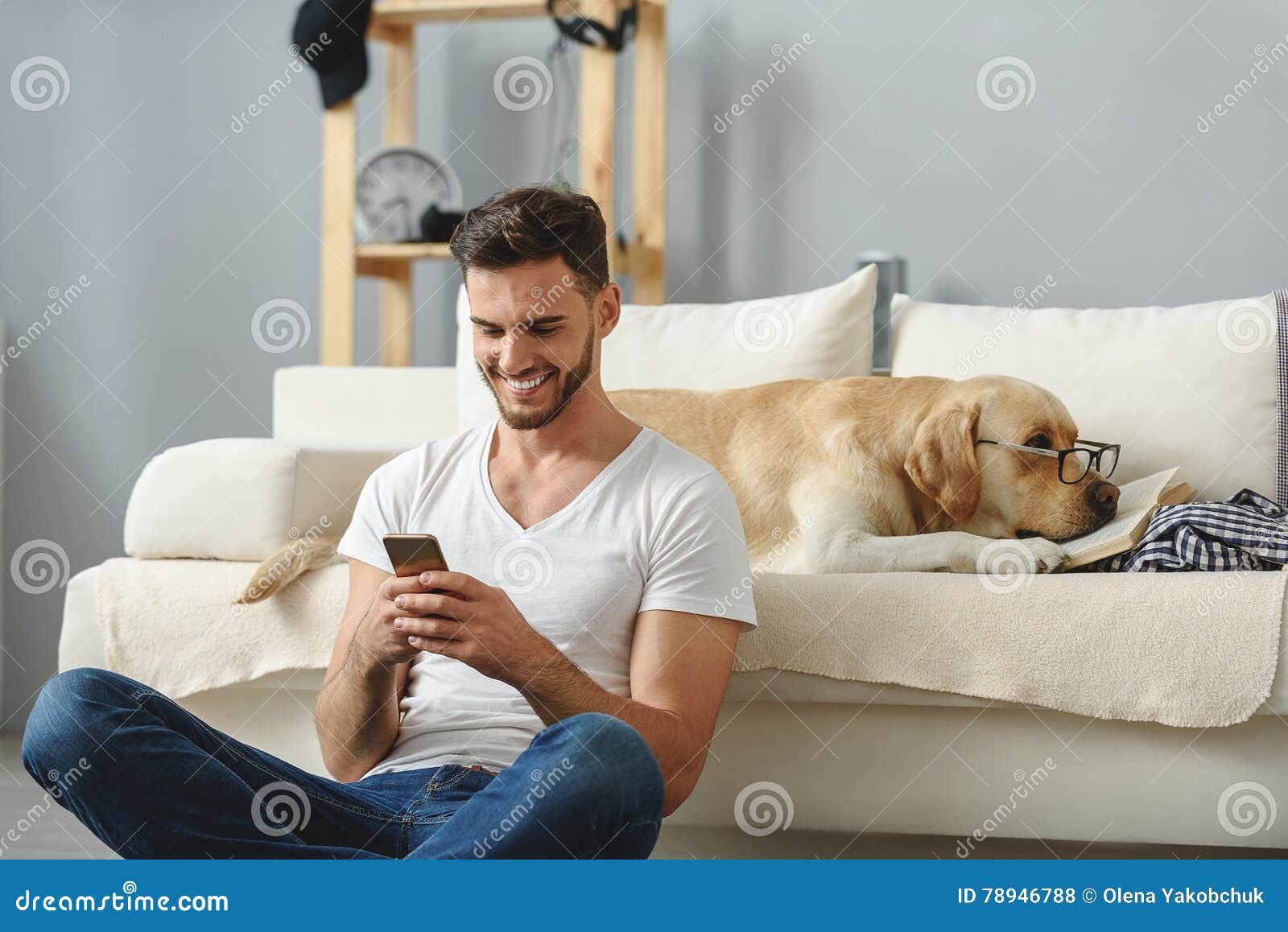 使用在智能手机的人，并且不注意他的狗. 英俊的人坐与盘的腿的地板有电话的，当说谎在沙发时的乏味小狗