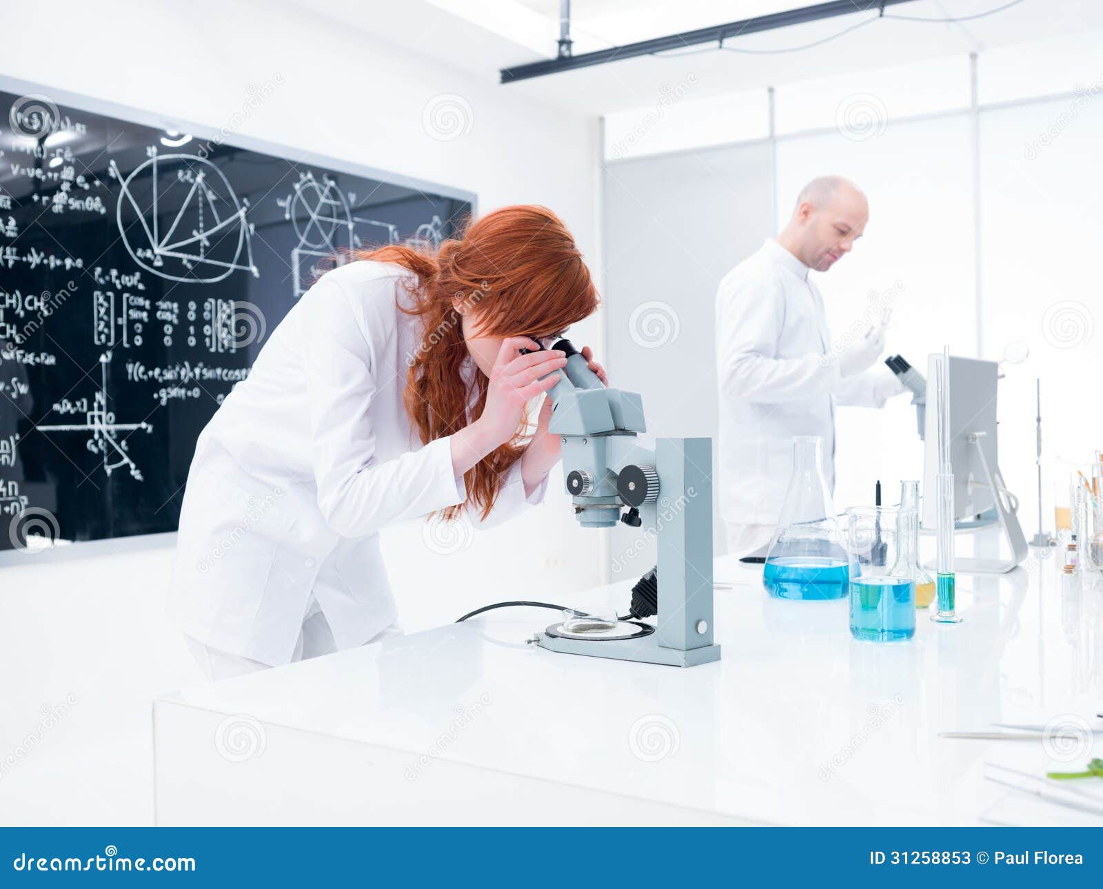 化学实验室实验. 分析在显微镜下的学生的全视图在监督在与实验室工具、五颜六色的液体和一个黑板的实验室桌附近的老师下在背景
