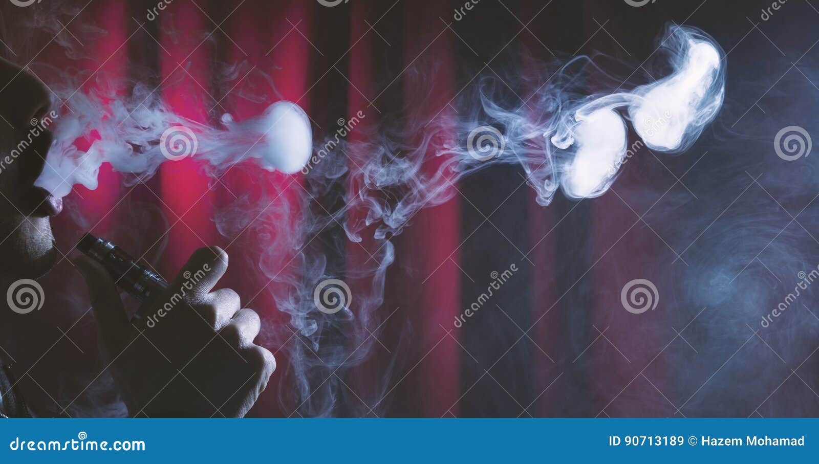 年轻在黑暗的背景的人vaping的云彩. 拿着电子香烟或e香烟与云彩的一个人在深红背景