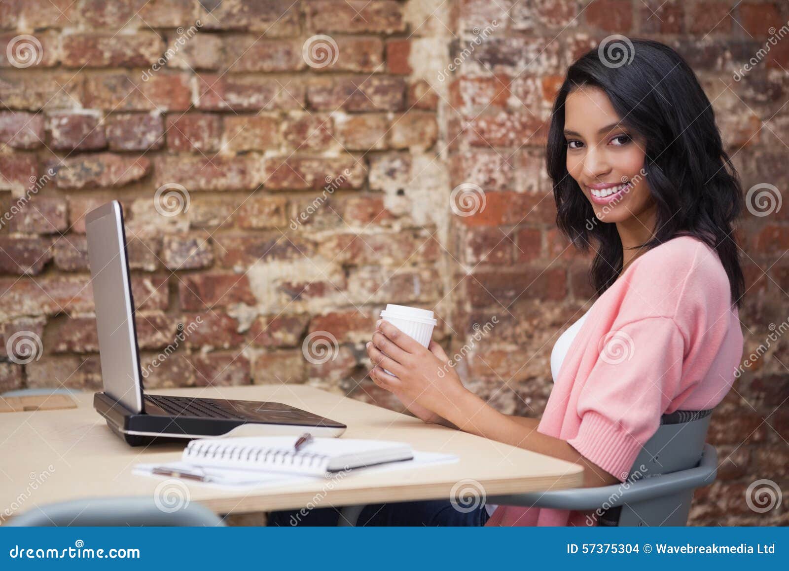 在她的书桌的微笑的妇女饮用的咖啡使用膝上型计算机在办公室