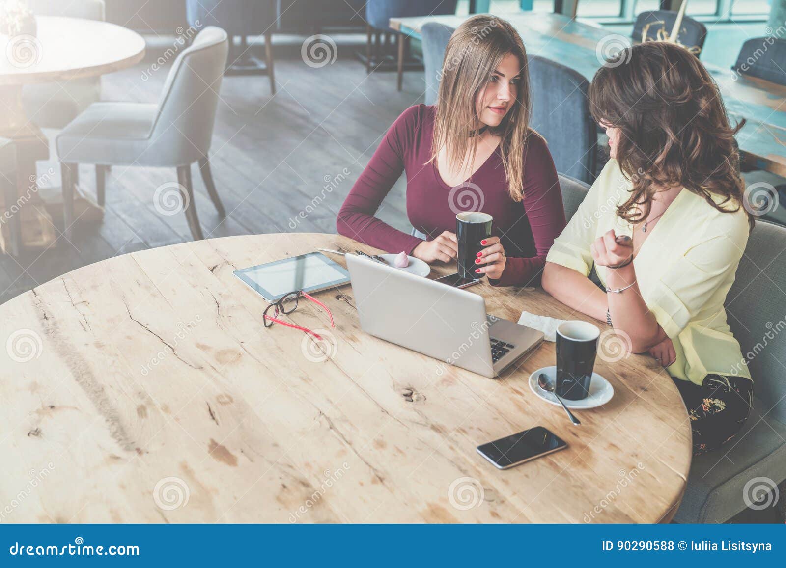 两名年轻可爱的妇女坐在咖啡馆，饮用的咖啡和谈话的圆的木桌上 在桌上是膝上型计算机、片剂计算机、智能手机和玻璃 业务会议，会议朋友，配合