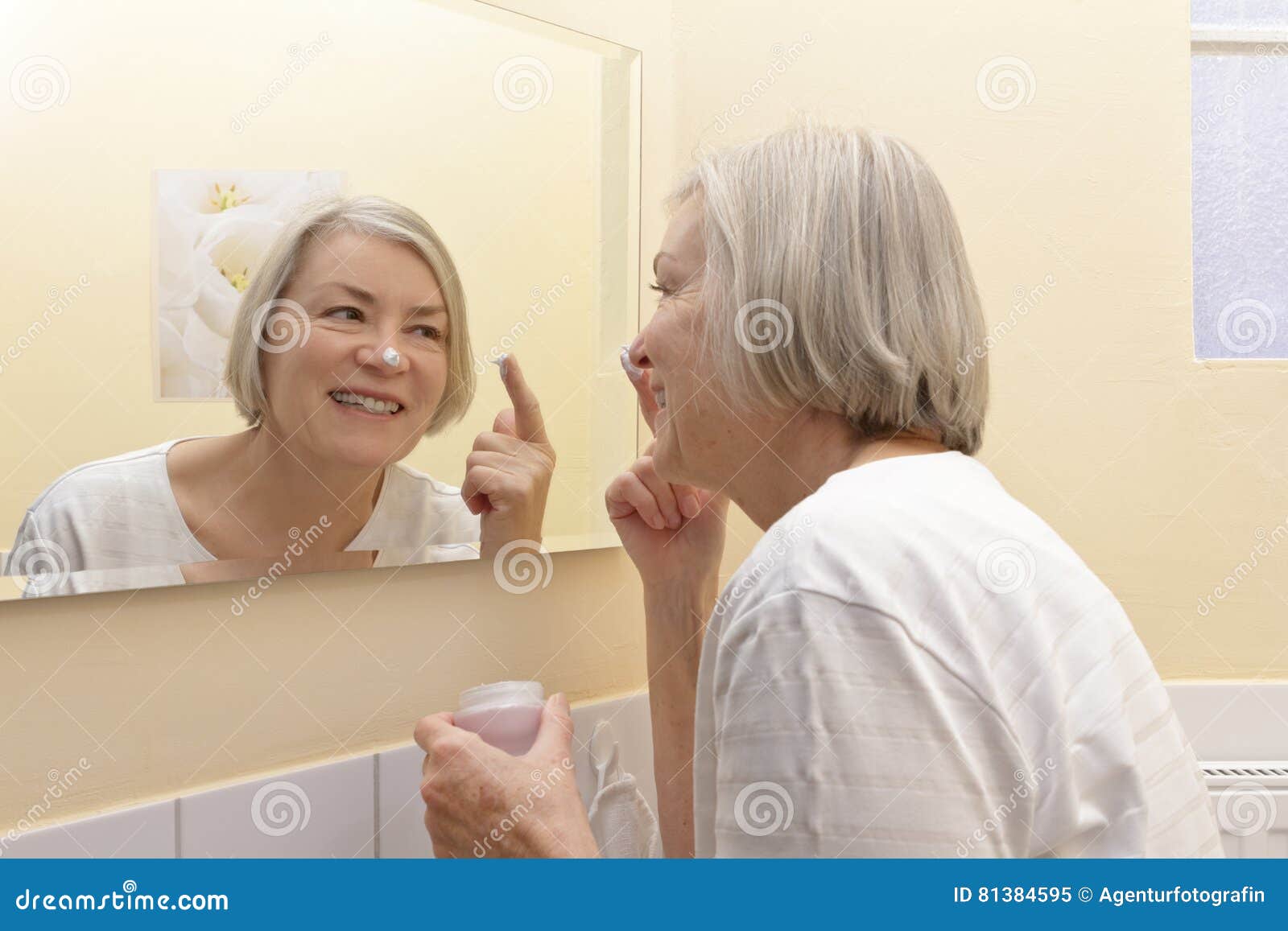 愉快的成熟妇女润肤霜镜子. 有一个奶油色瓶子的快乐的资深妇女在她的手上和在她的站立在应用润肤霜的她的黄色卫生间前面镜子的鼻子的一个白色斑点于她的面孔