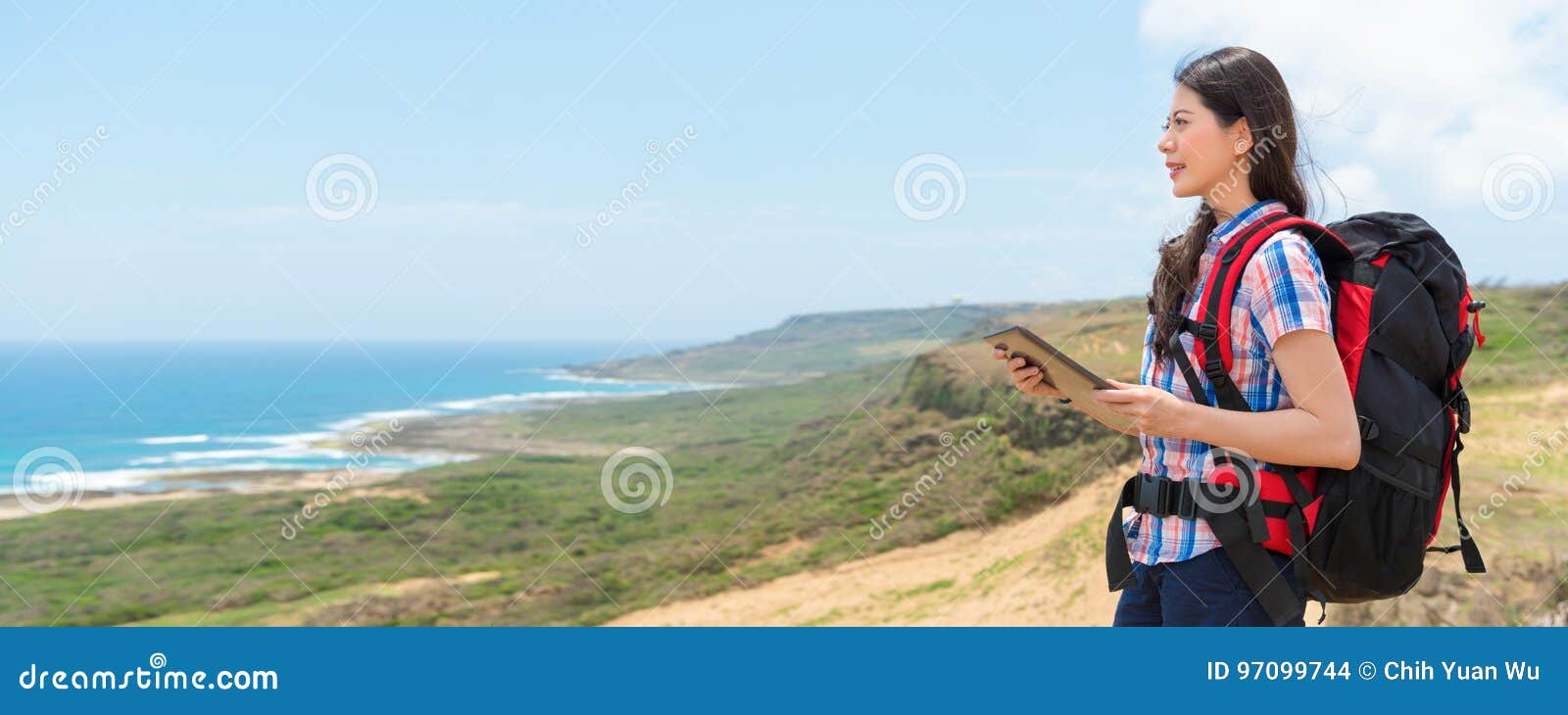 拿着网上旅行指南的女性背包徒步旅行者. 拿着网上旅行指南的微笑的中国女性背包徒步旅行者通过数字式片剂填塞在远足享受与横幅庄稼的山的身分海洋风景拷贝空间的