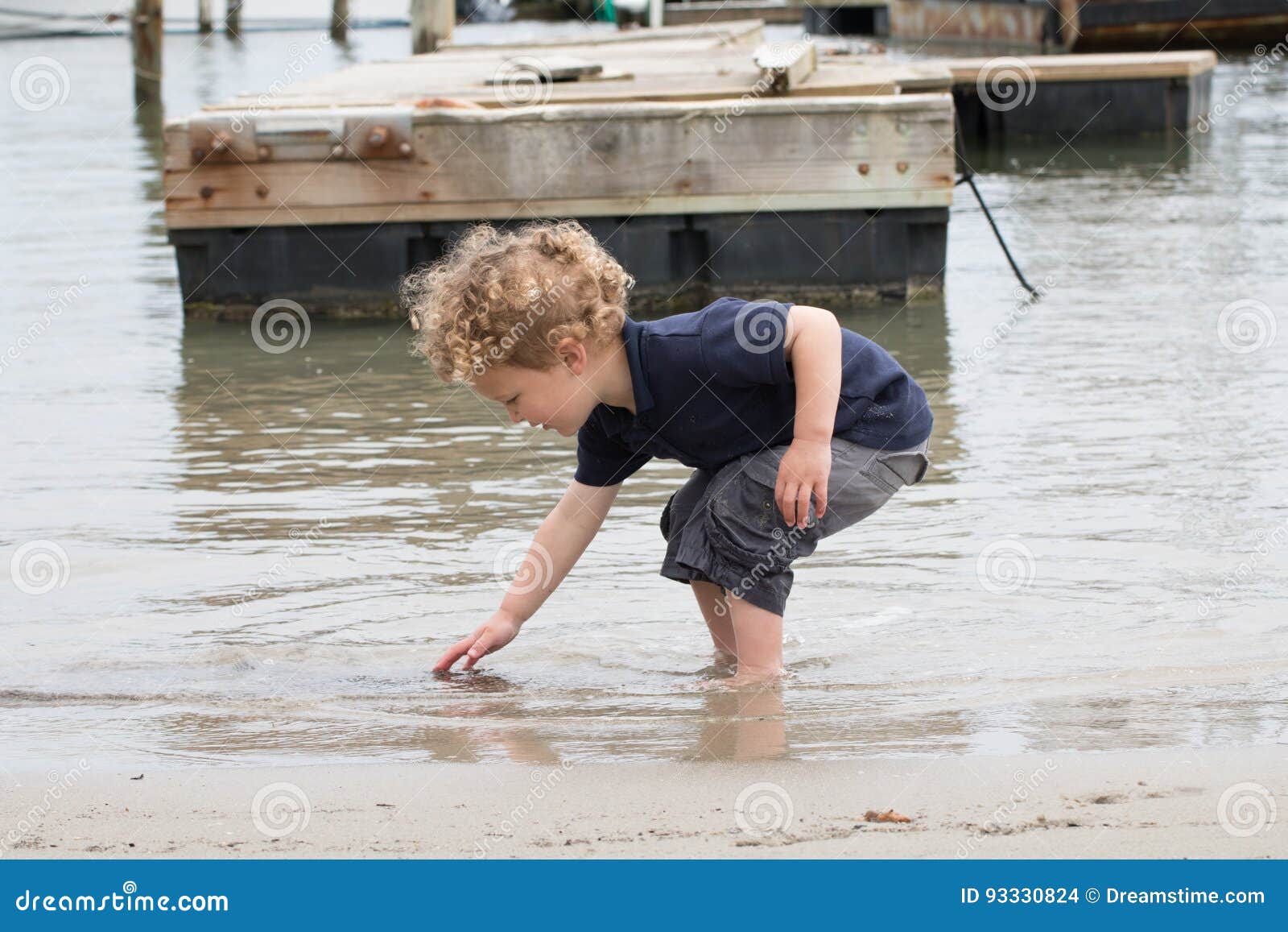 搜寻壳的年轻男孩在港口. 一个幼儿在罗德岛州渔村寻找壳或岩石 有一个老船坞在背景中 男孩有白肤金发的卷发并且是大约3岁
