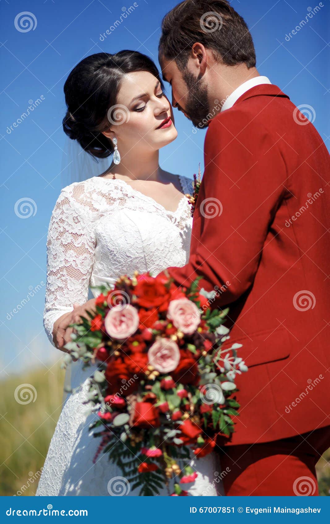 新娘和新郎，室外可爱的夫妇，与红色花的婚姻的新娘花束 蓝天，绿草在背景中. 新娘和新郎，室外可爱的夫妇，巨大别致的与红色花的婚礼新娘花束 蓝色草绿色天空 夫妇的垂直的秀丽画象 有胡子和髭的时髦的人在红色衣服 婚礼颜色马尔萨拉