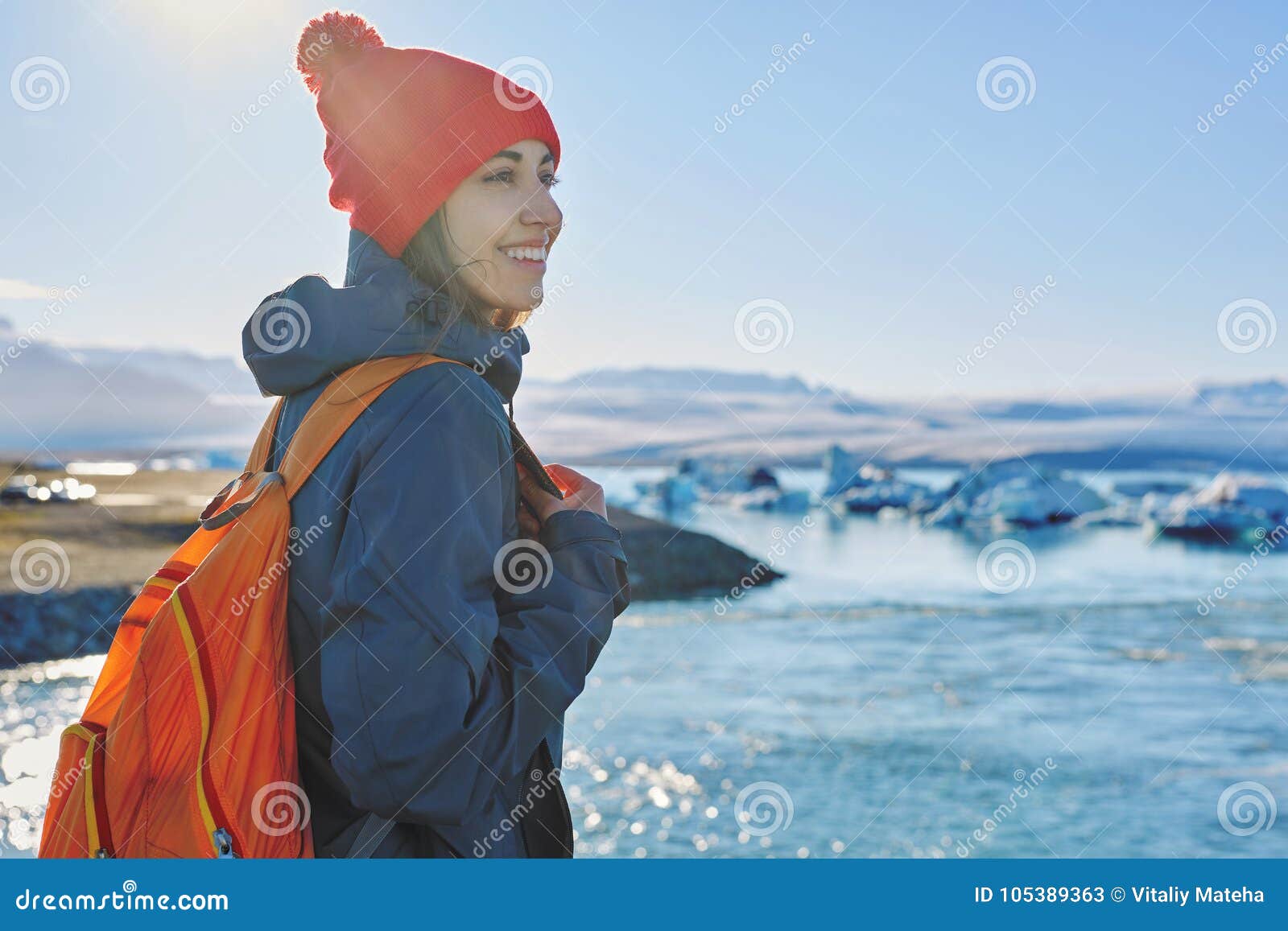 有背包的妇女在冰盐水湖在冰岛. 女孩画象温暖的衣物的有橙色背包的在冰盐水湖在冰岛有海和天空背景