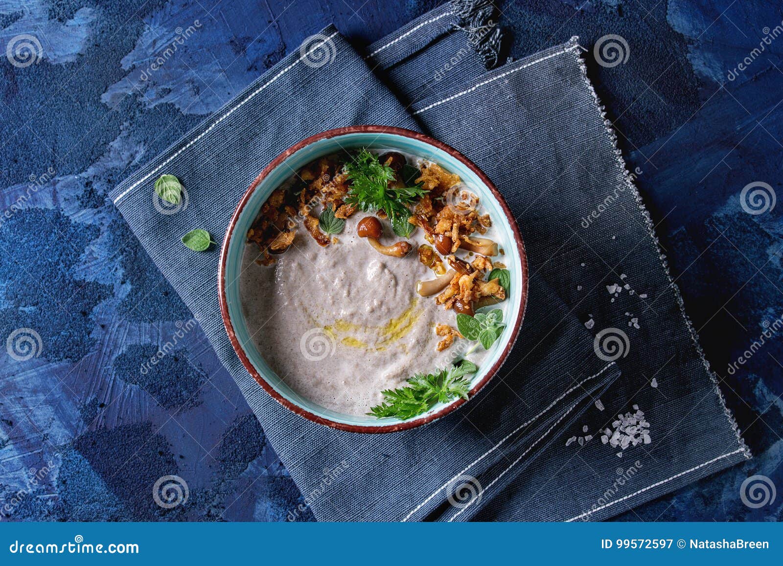 在陶瓷碗的蘑菇奶油色汤服务用森林蘑菇，绿色，在蓝色纺织品餐巾的油煎的葱在深蓝纹理混凝土背景 与空间的顶视图